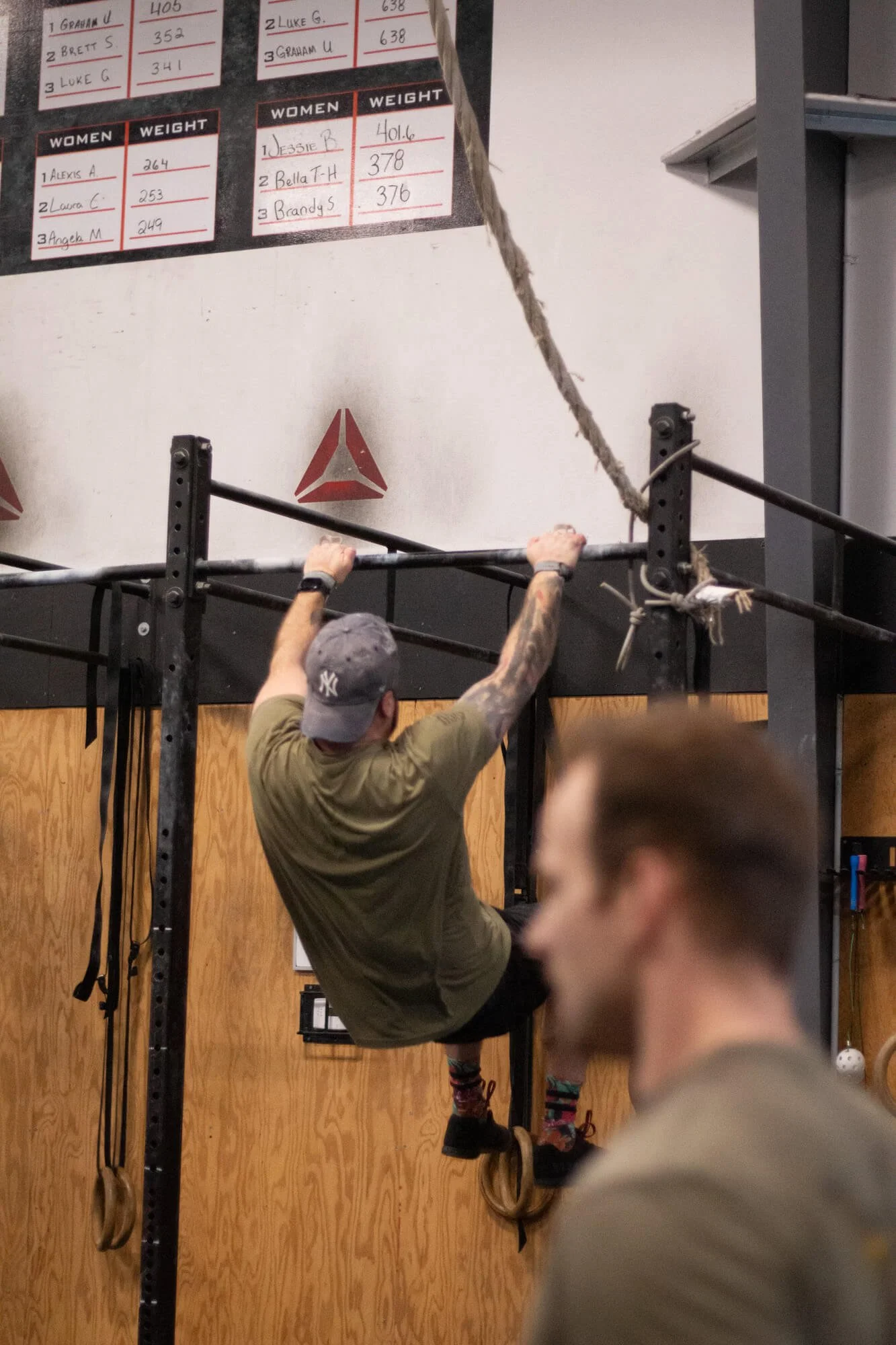 A man in a gray cap and green t-shirt is climbing gymnastic rings at a gym, hanging from a climbing rope. A blurred person is in the foreground. A scoreboard with workout scores is on the wall behind them.