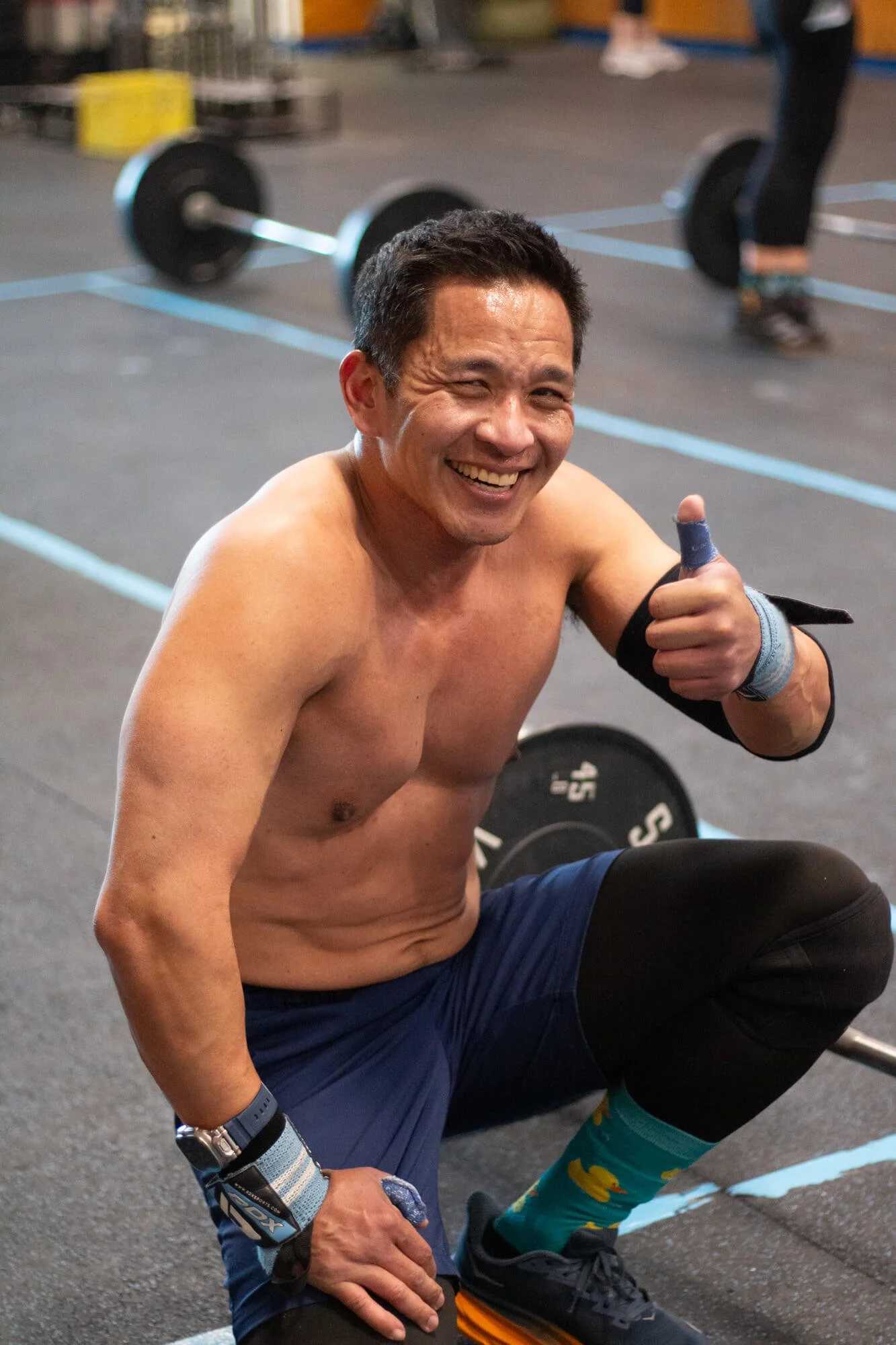 A smiling man with short dark hair, shirtless, wearing workout gear including knee sleeves, wrist wraps, and colorful socks, sitting on the floor at a gym, giving a thumbs-up, with barbells and weight plates visible in the background.
