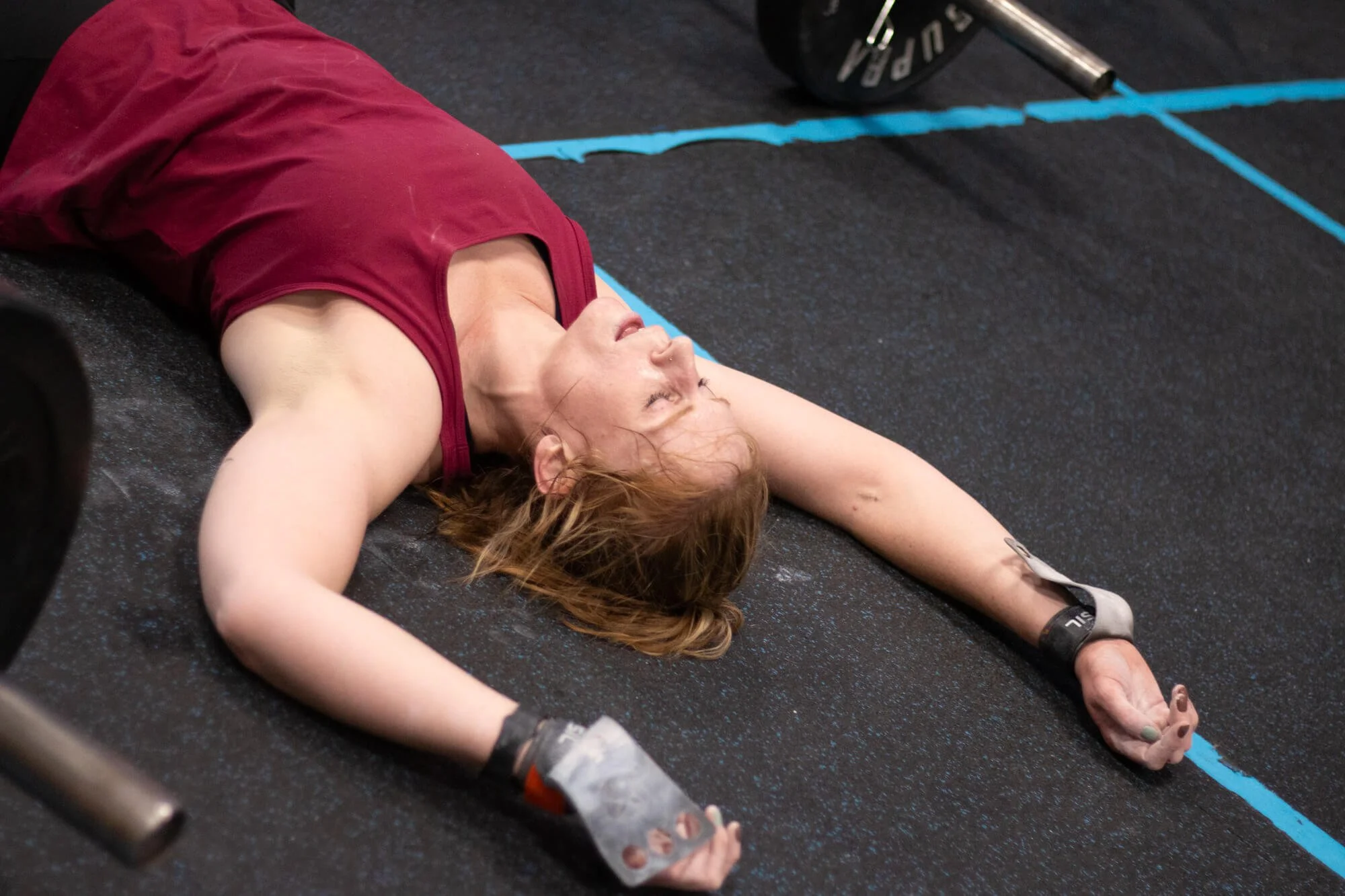 Woman lying on the gym floor after a workout, wearing workout gloves and a maroon tank top.
