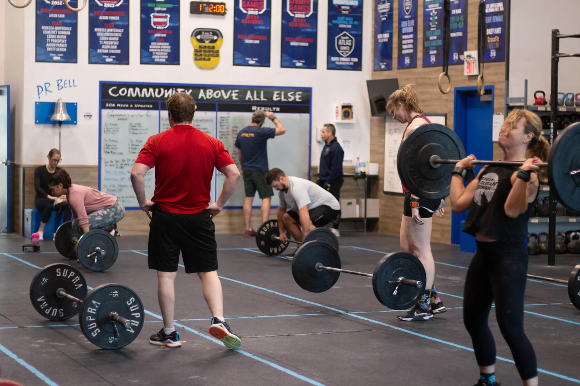 People lifting weights in a gym with a coach observing and a scoreboard on the wall.