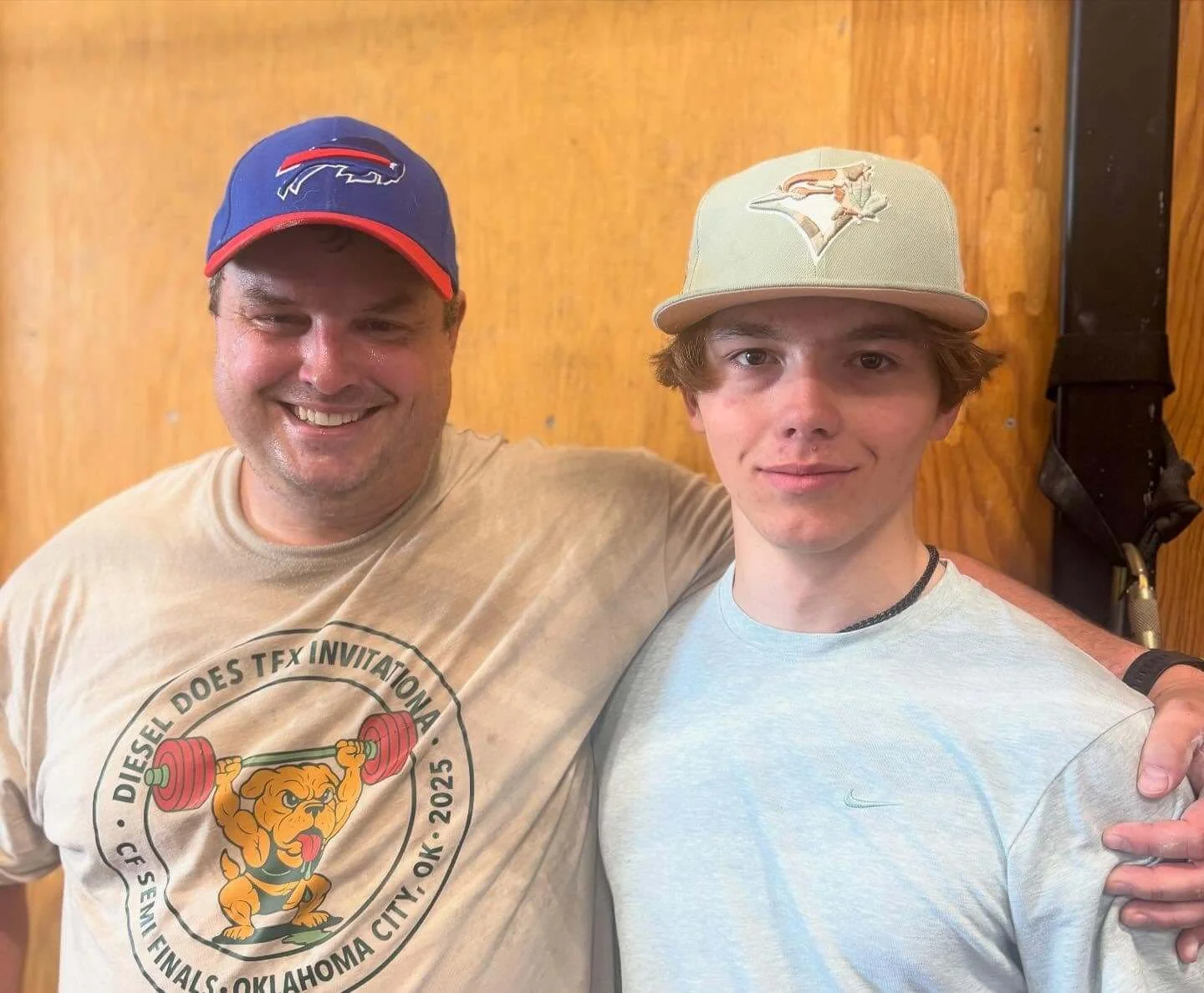 Two men standing close together in front of a wooden wall, smiling and looking at the camera. The man on the left is wearing a blue and red baseball cap with a sports logo, a beige T-shirt with a lion mascot lifting weights, and has his arm around the shoulder of the man on the right. The man on the right is wearing a light gray Nike T-shirt and a beige baseball cap with a logo of a bird.
