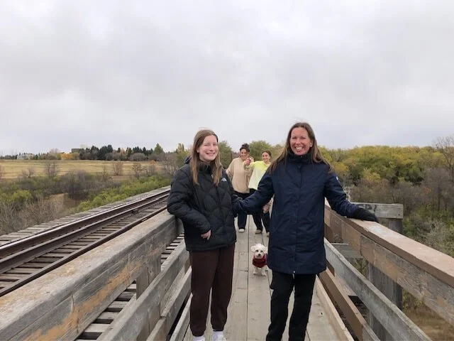 Group of people walking on a wooden bridge near railroad tracks, smiling, one person holding a dog, overlooking a landscape with trees and fields under a cloudy sky.