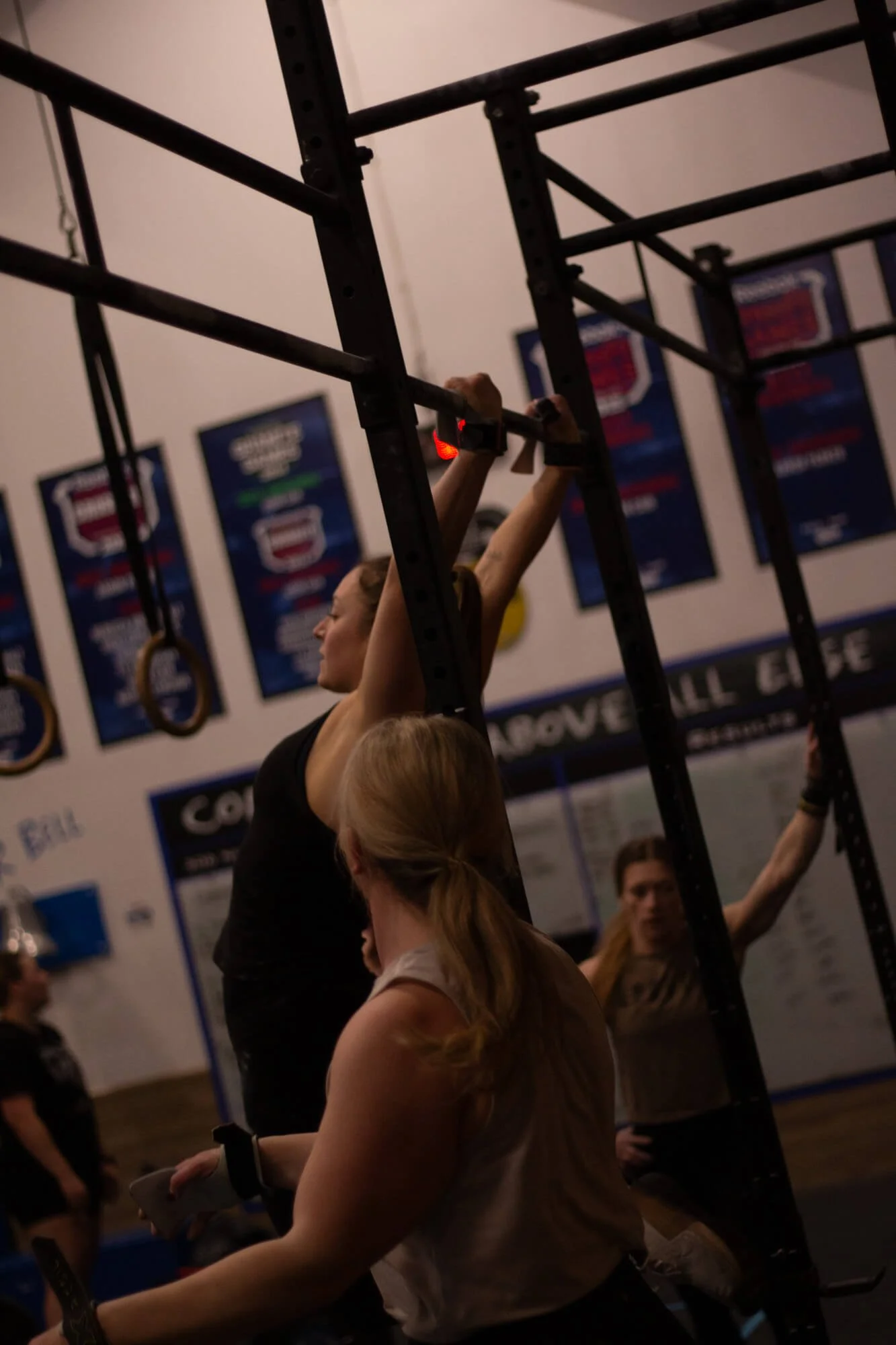 A woman with blonde hair in a ponytail assisting a woman lifting her arms during a workout at a gym, with people and gym banners in the background.