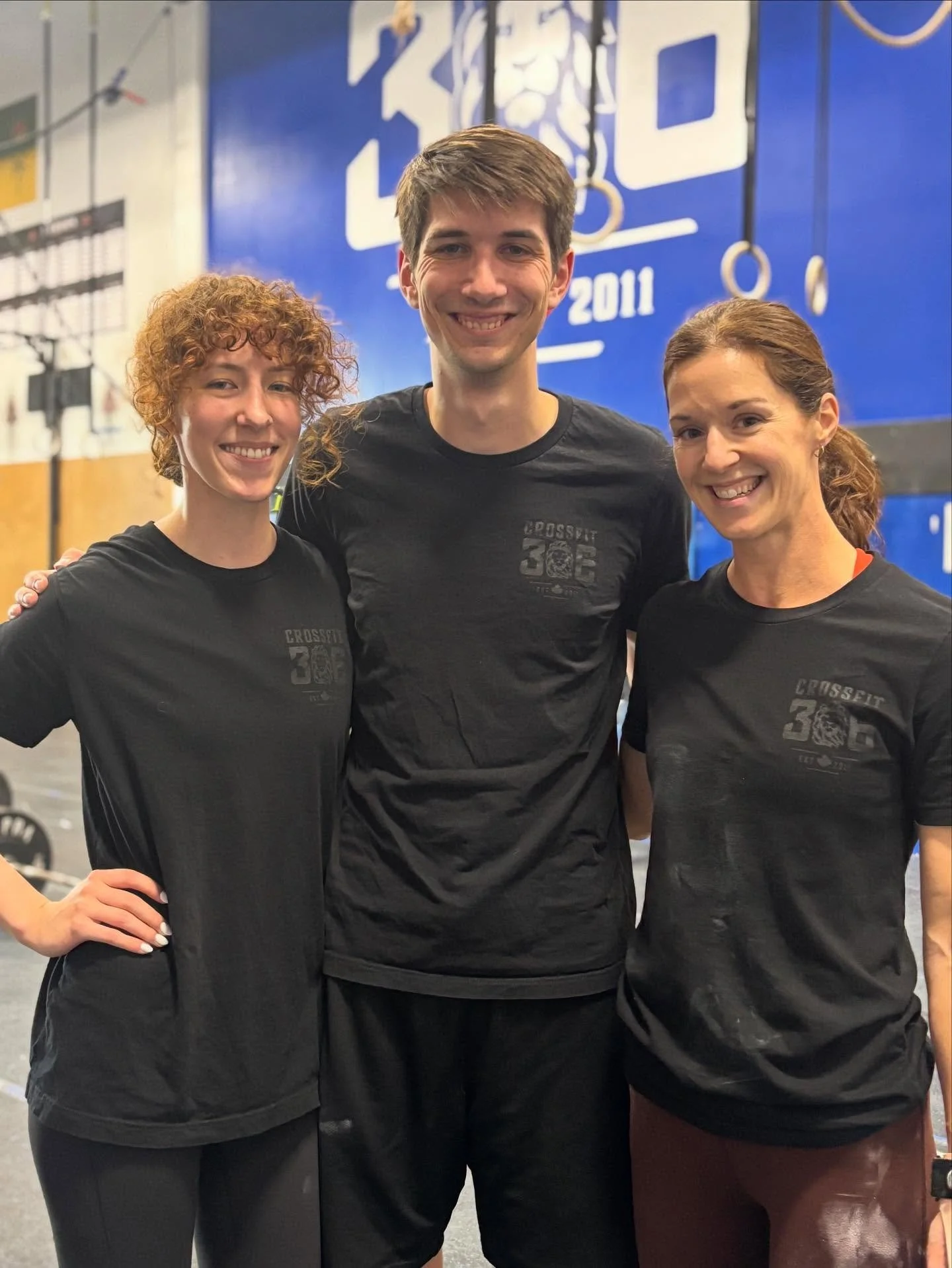Three smiling people wearing black CrossFit shirts, standing together in a gym. The background shows CrossFit equipment and murals.