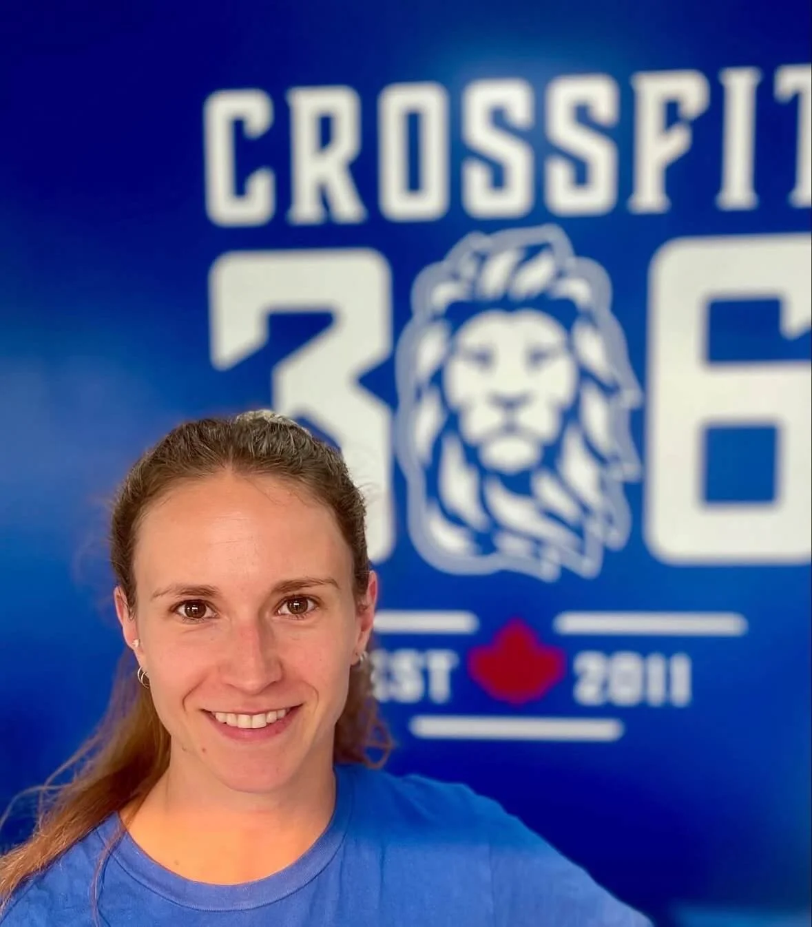 A woman taking a selfie in front of a blue CrossFit 306 logo with a lion's head. She has long brown hair, is smiling, and wearing a blue shirt.