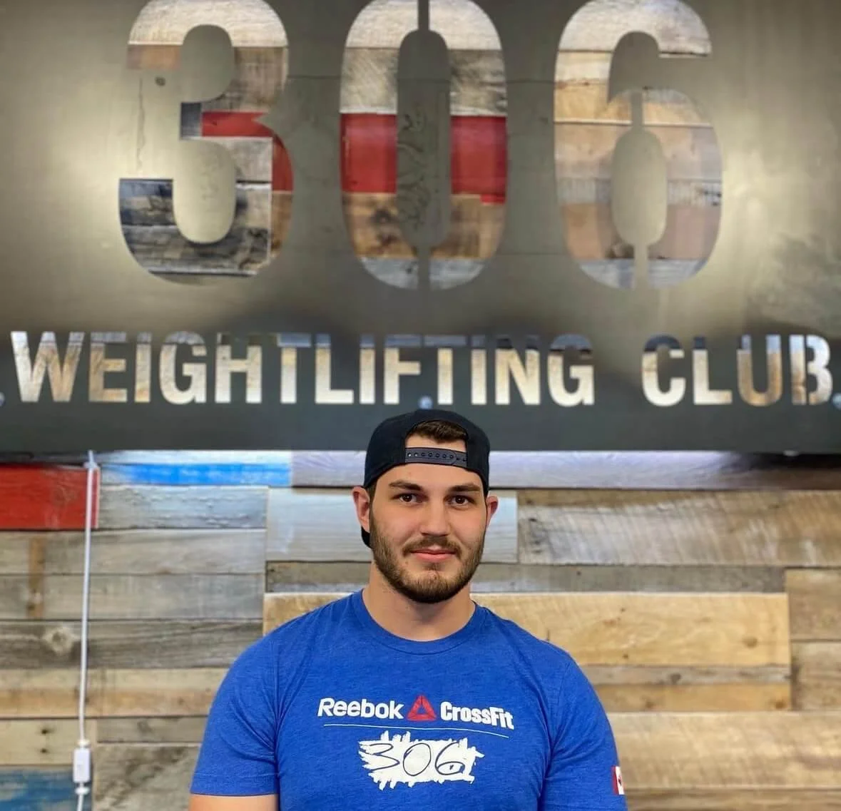 A man standing in front of a sign that reads '306 WEIGHTLIFTING CLUB'. He is wearing a blue Reebok CrossFit t-shirt and a black baseball cap worn backwards.