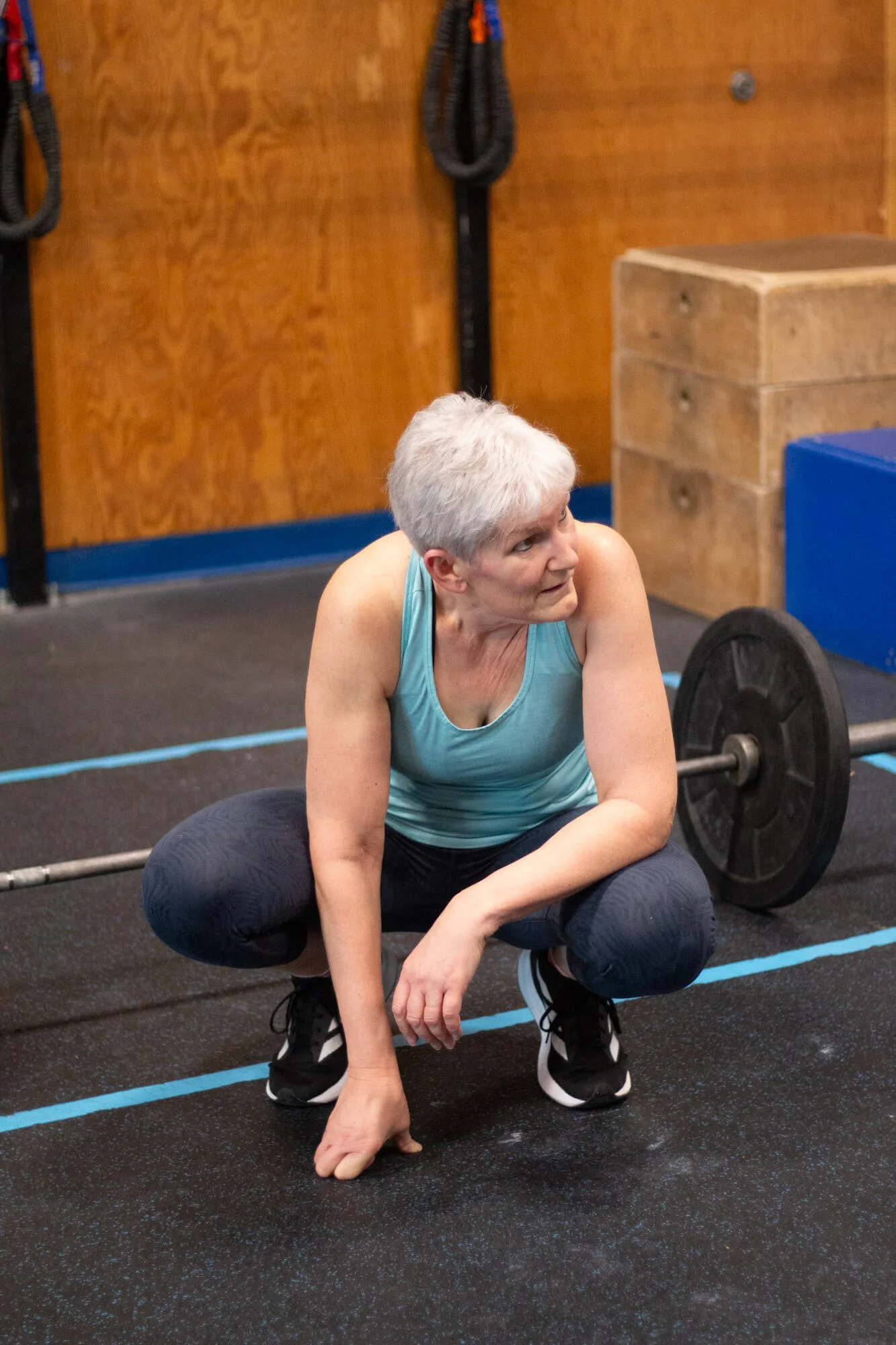 An older woman with gray hair in a gym, squatting on the floor next to a barbell, wearing a light blue athletic tank top and dark leggings.