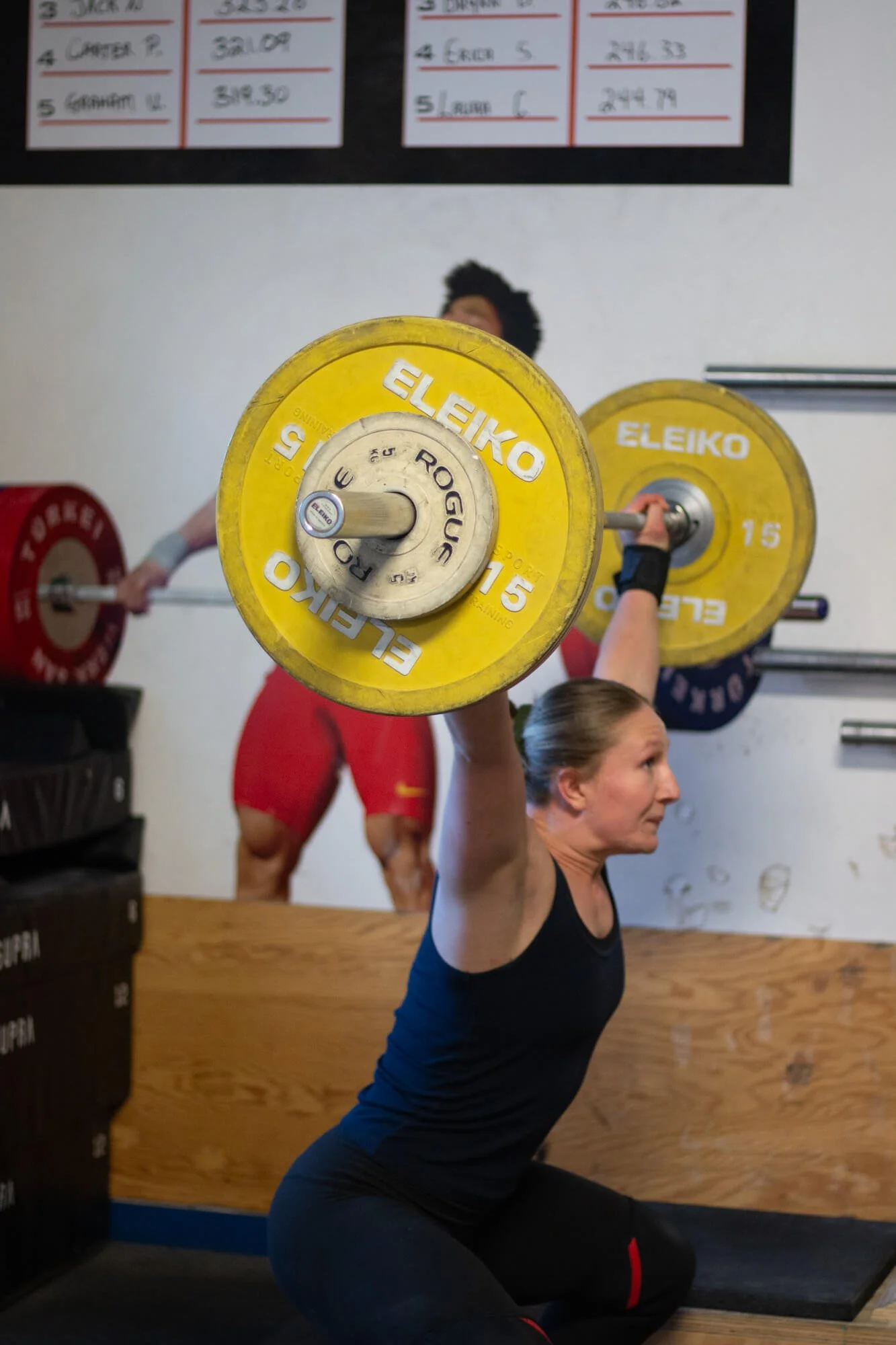 A woman lifting a barbell with yellow and black weights during a weightlifting exercise at a gym. A man is in the background, also lifting weights.