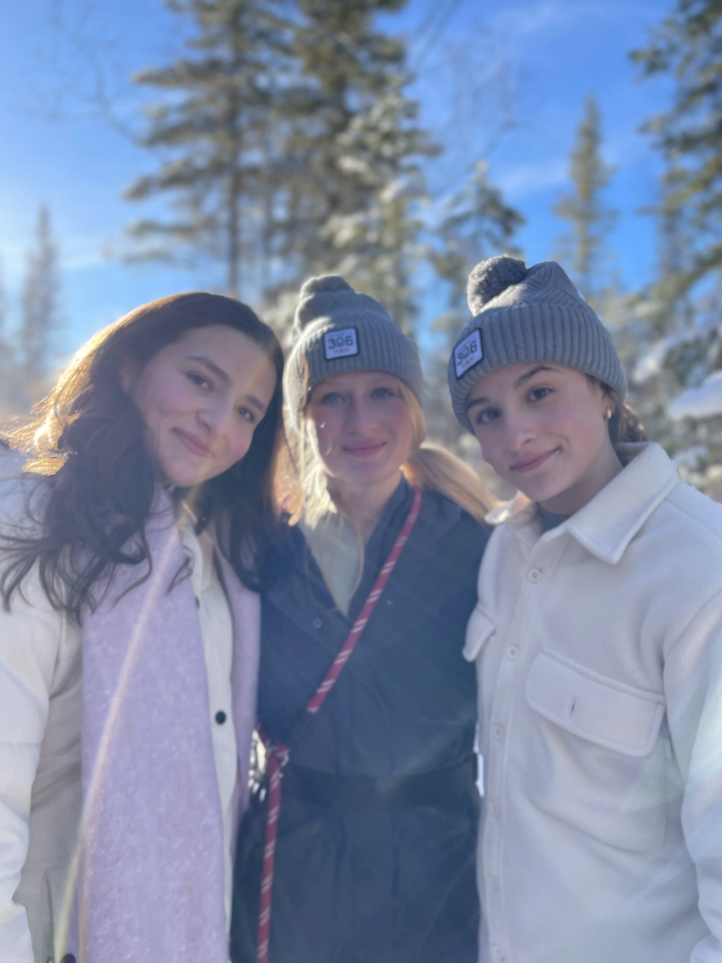 Three young women standing outdoors in a snowy forest, wearing winter clothing and grey beanies, smiling at the camera.