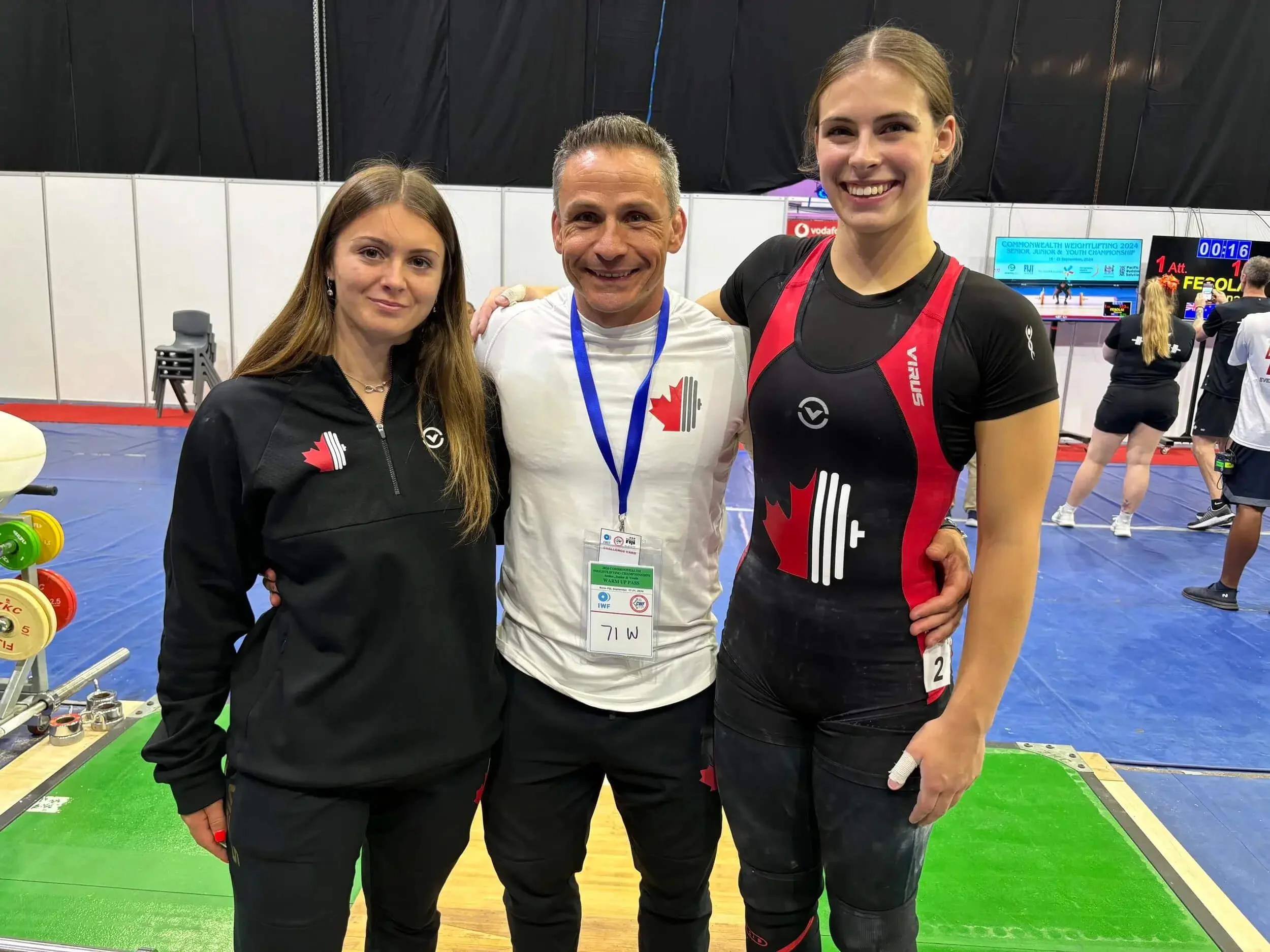 Three people, a woman in black athletic jacket, a man in white shirt with Canadian flag, and a tall woman in a black and red athletic uniform, posing inside a sport hall with weightlifting equipment and other athletes in the background.
