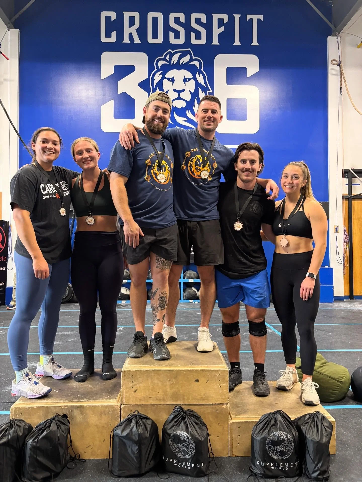 Group of six athletes standing on a wooden podium at a CrossFit competition, smiling and wearing medals, with a large CrossFit 306 sign and lion logo in the background.