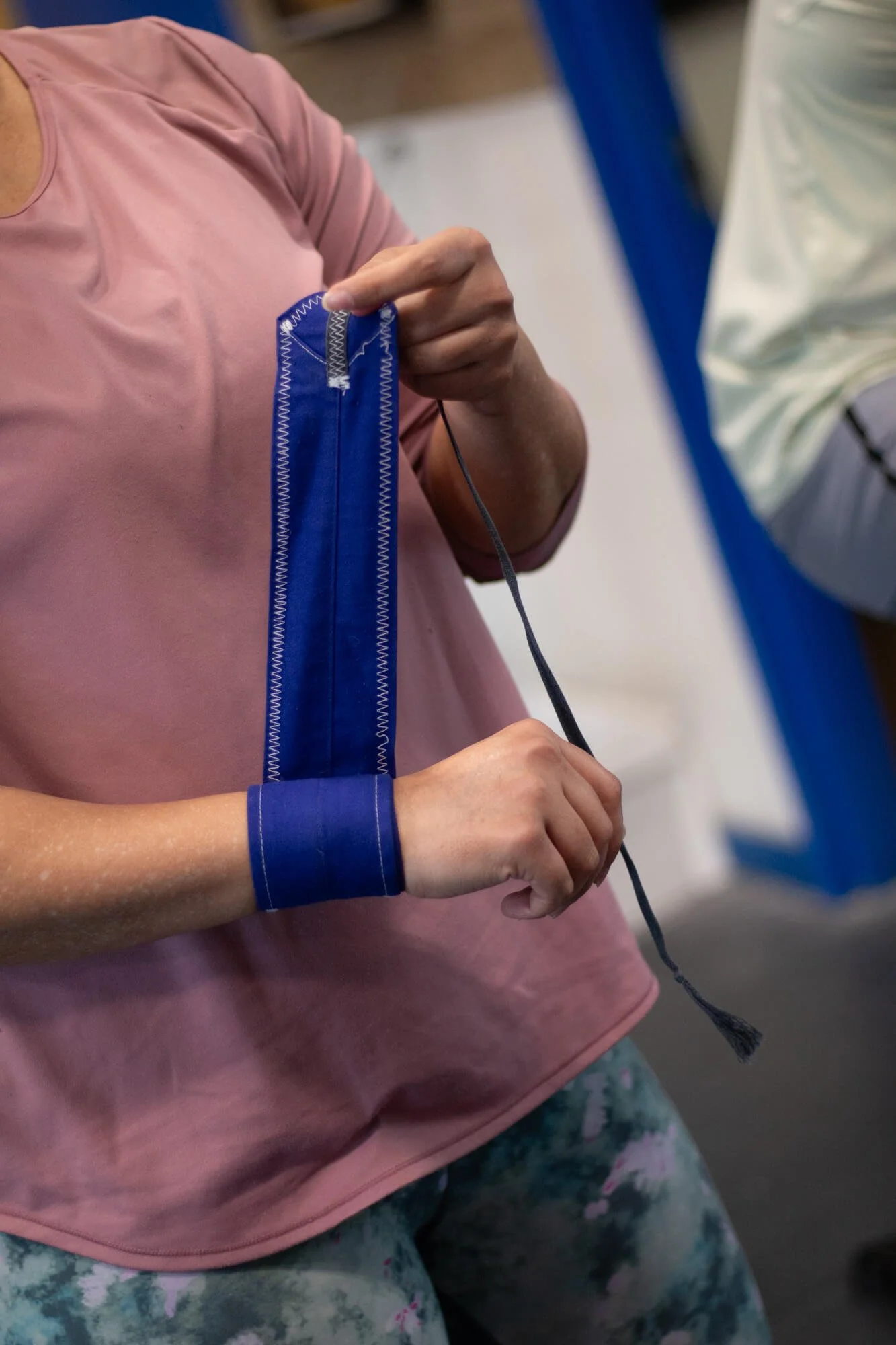 A person wearing a pink shirt and patterned leggings tying a blue wristband, with another person nearby.