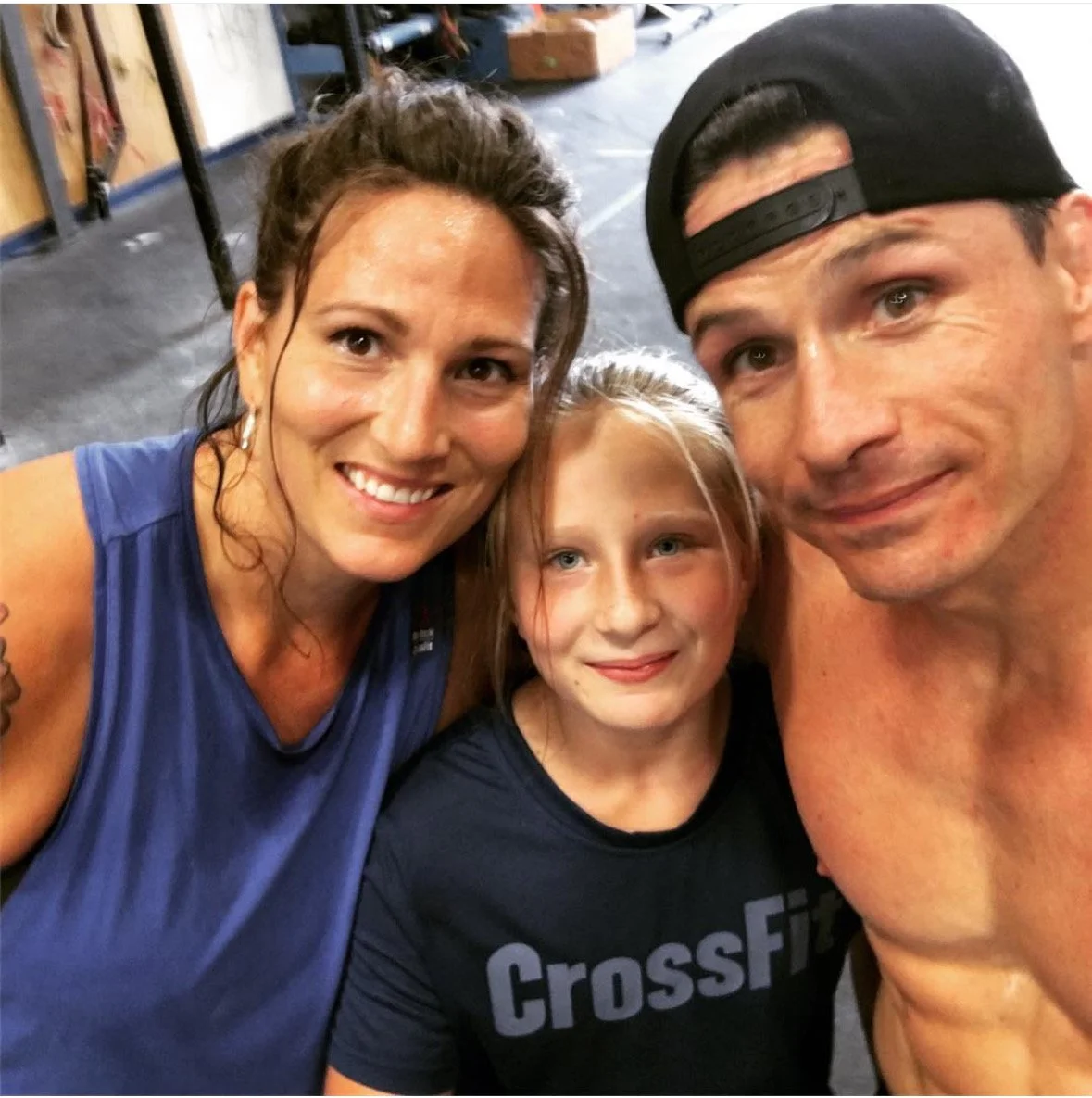 A family of three taking a selfie after a workout at the gym, with the mother on the left, the daughter in the middle wearing a CrossFit t-shirt, and the father on the right wearing a backwards cap.