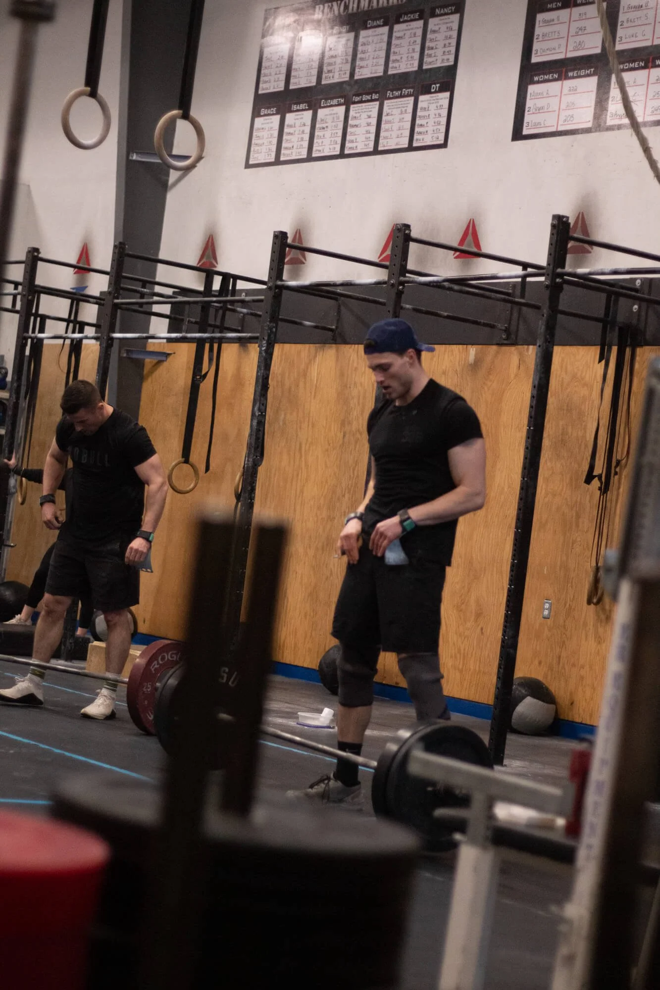 Two men in a gym, one lifting a barbell with weights and the other standing nearby, with gym equipment and fitness charts on the wall in the background.