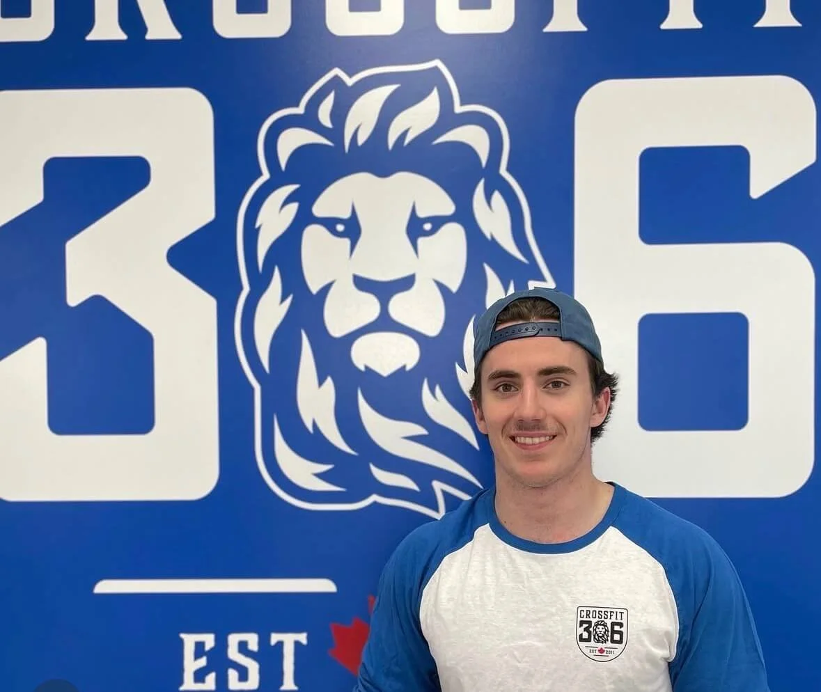 A young man with dark hair, wearing a blue baseball cap backward and a white and blue CrossFit shirt, smiling in front of a large blue wall with a white lion logo and the text '306'