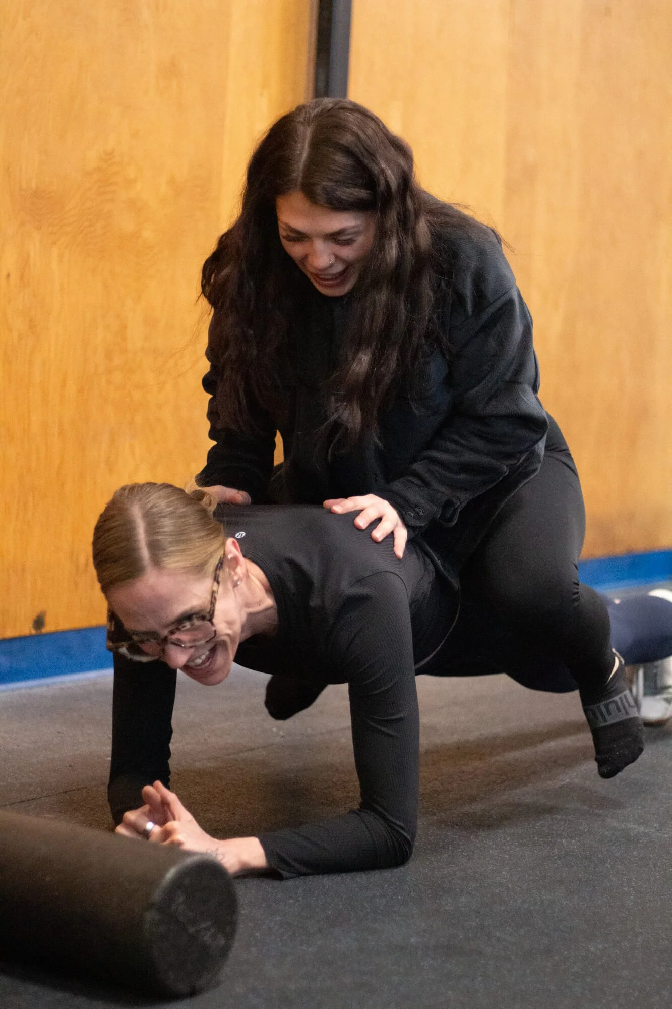 Two women are engaging in a playful moment, with one bending over and the other sitting on her back, both smiling and laughing indoors.