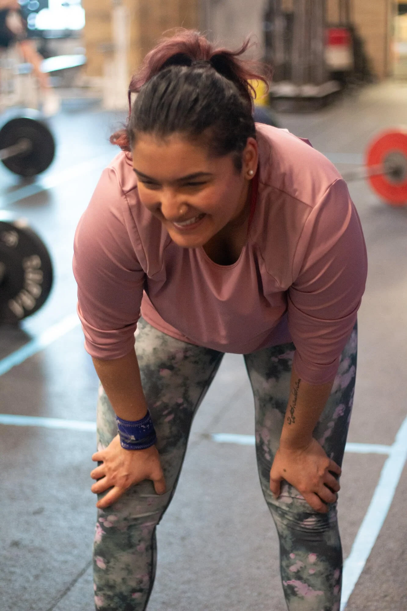 A woman with dark hair in a ponytail, wearing a pink shirt and patterned leggings, smiling while leaning forward with hands on her knees inside a gym.