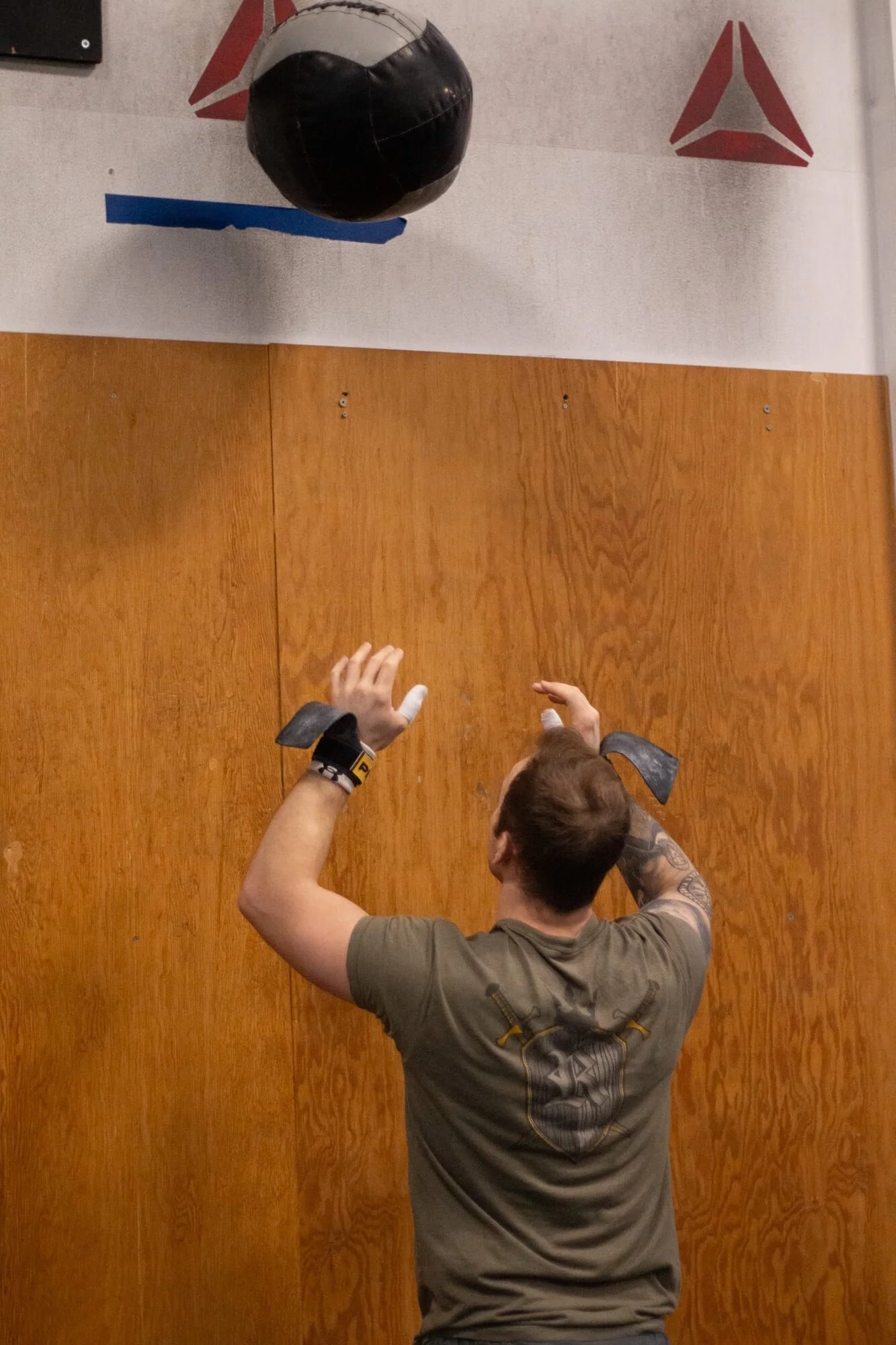 A person with tattoos and a brown T-shirt is practicing wall climbing with a black medicine ball hanging above them inside a gym. There is a wooden wall in the background.