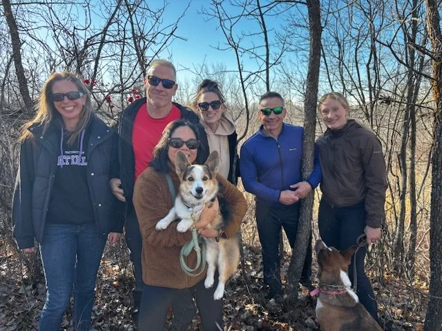 Six people pose outdoors in a wooded area on a clear day, some wearing sunglasses, with two dogs among them.