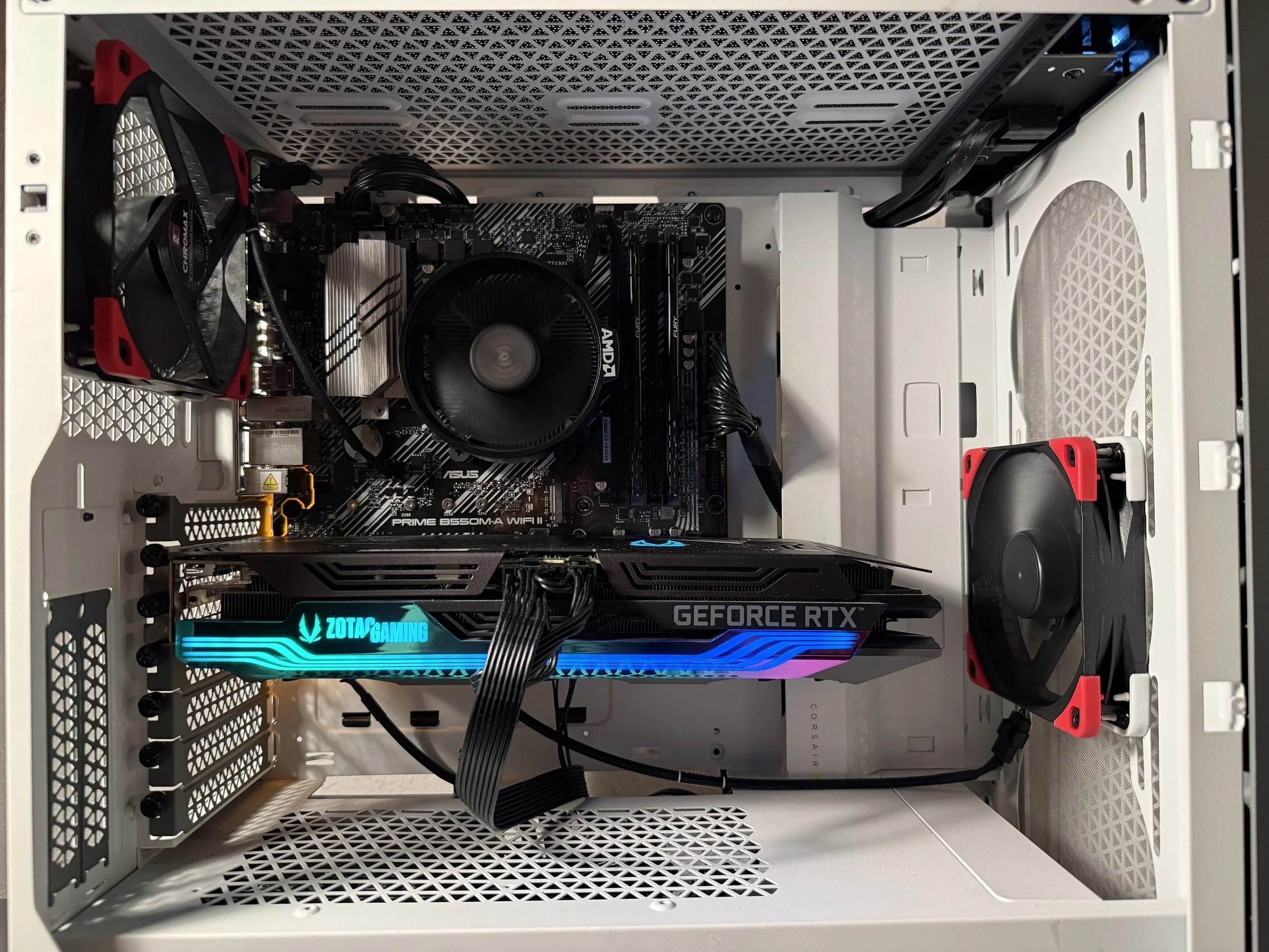 Inside a white computer case with black and red cooling fans, a black and silver Asus motherboard, a black AMD CPU cooler, a Zotac Gaming GeForce RTX graphics card with RGB lighting, and black power supply cables.