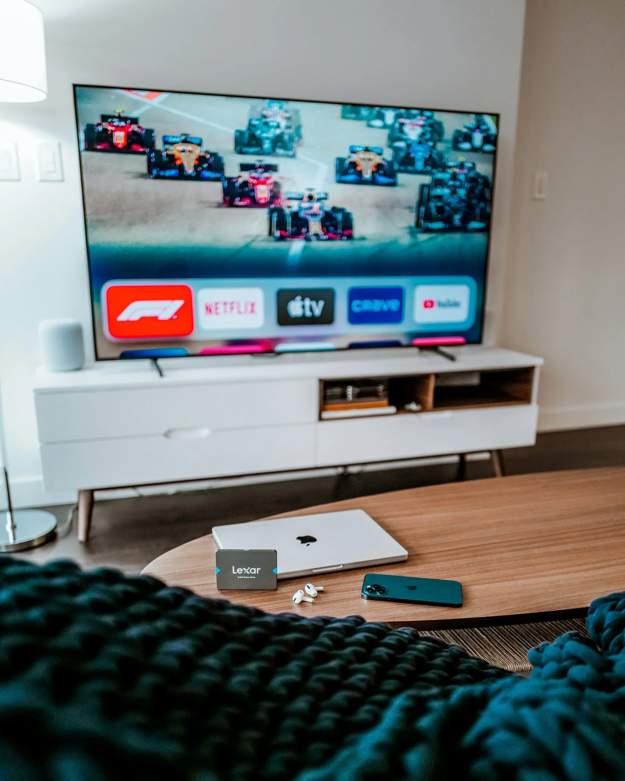 Living room with a TV displaying a Formula 1 race, placed on a white TV stand. On the wooden coffee table, there is a closed MacBook, wireless earbuds, a Lenovo computer card, and an iPhone. Part of a knitted blanket is visible in the foreground.