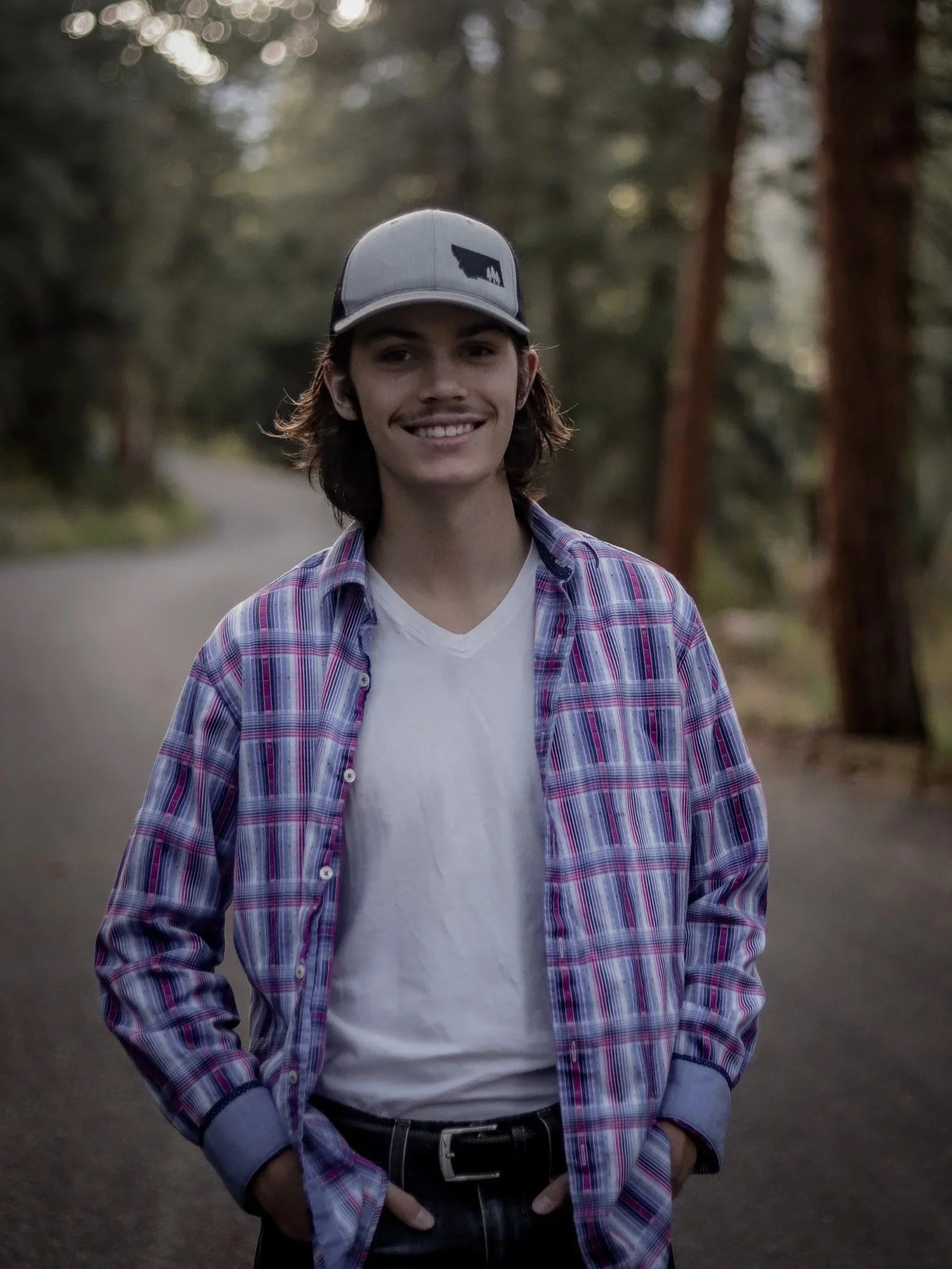 A young man smiling, standing outdoors on a forest path, wearing a baseball cap, plaid shirt, and white t-shirt.