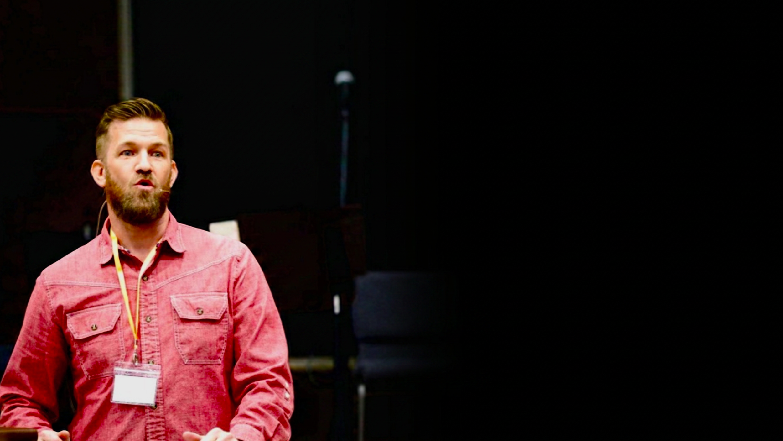 A man with a beard and short hair wearing a pinkish-red shirt standing against a dark background.