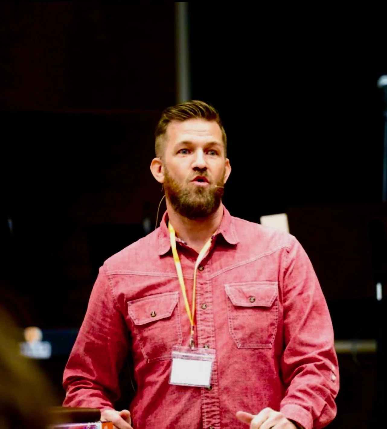 A man with a beard and short hair wearing a pink button-up shirt, speaking at an event with a lanyard and badge around his neck, in front of dark background.