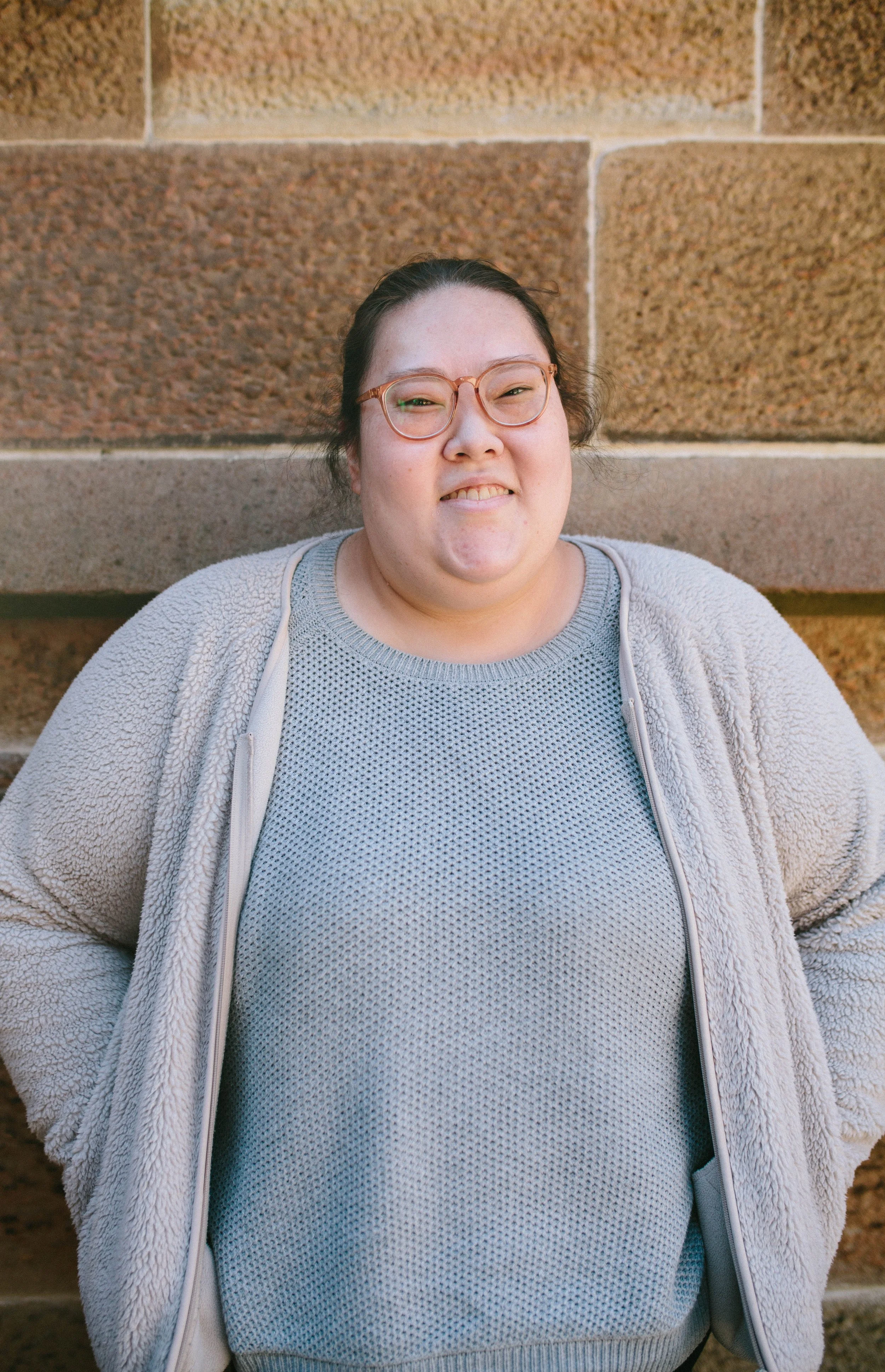 A woman with glasses smiling, standing against a brown stone wall.