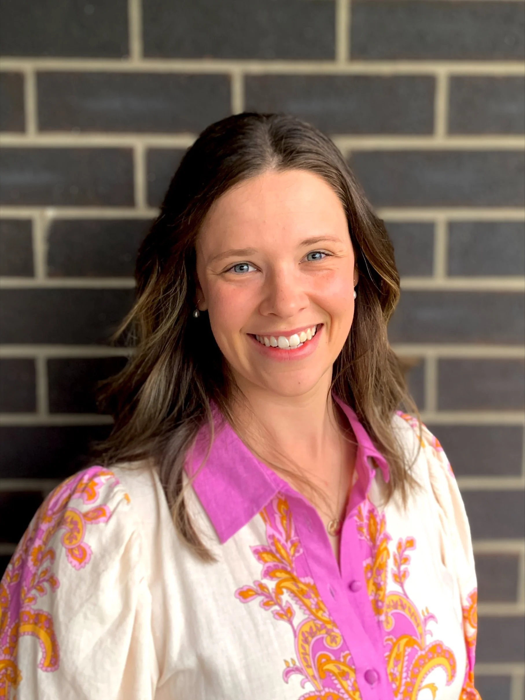 A woman smiling with a black brick wall background, wearing a cream-colored blouse with pink and yellow embroidery and a pink collar.