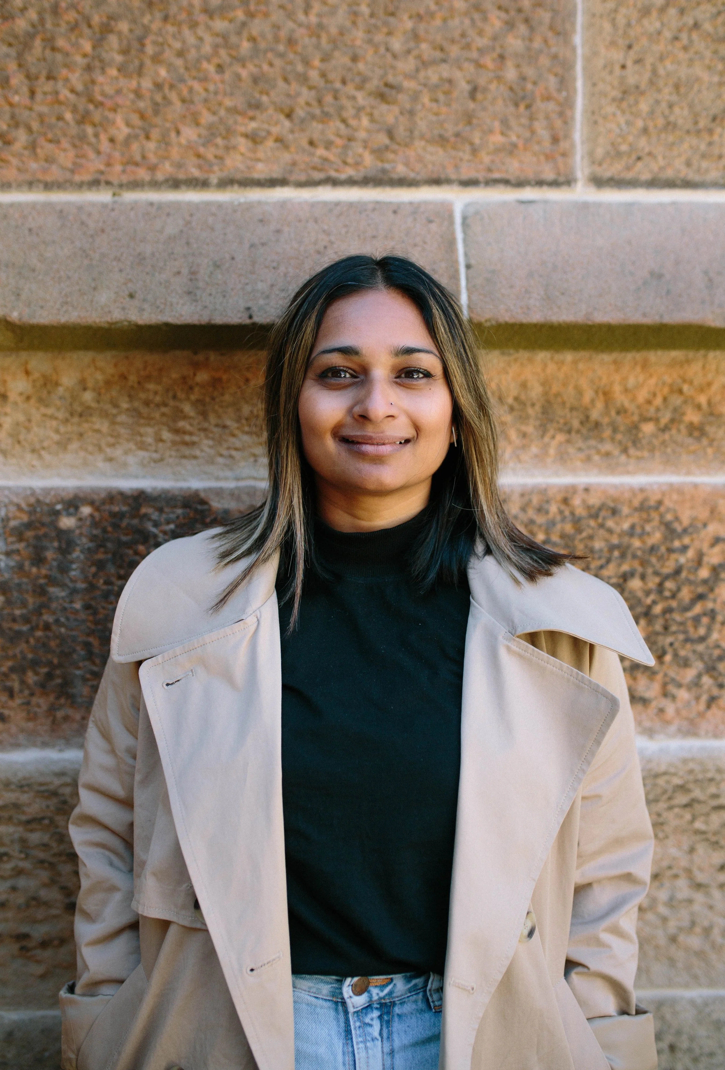 A woman with shoulder-length dark hair and light brown highlights, wearing a beige trench coat over a black turtleneck, standing against a textured brick wall, smiling gently.