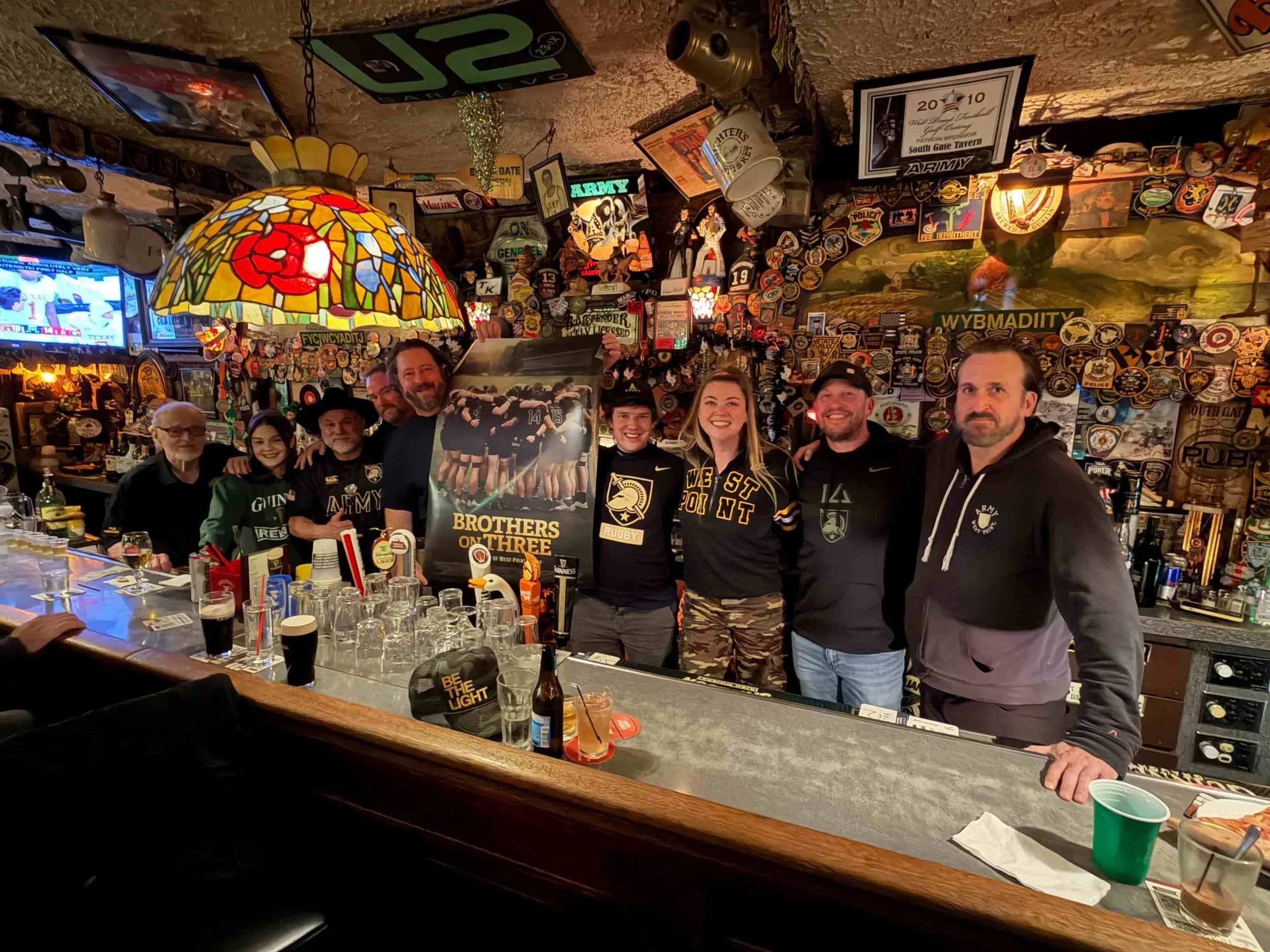A group of seven people, four men and three women, standing behind a bar in a decorated pub with trophies, patches, and signs, including a neon army sign. They are smiling and holding a framed picture of a rugby team, with various bar items and glasses on the counter in front of them.