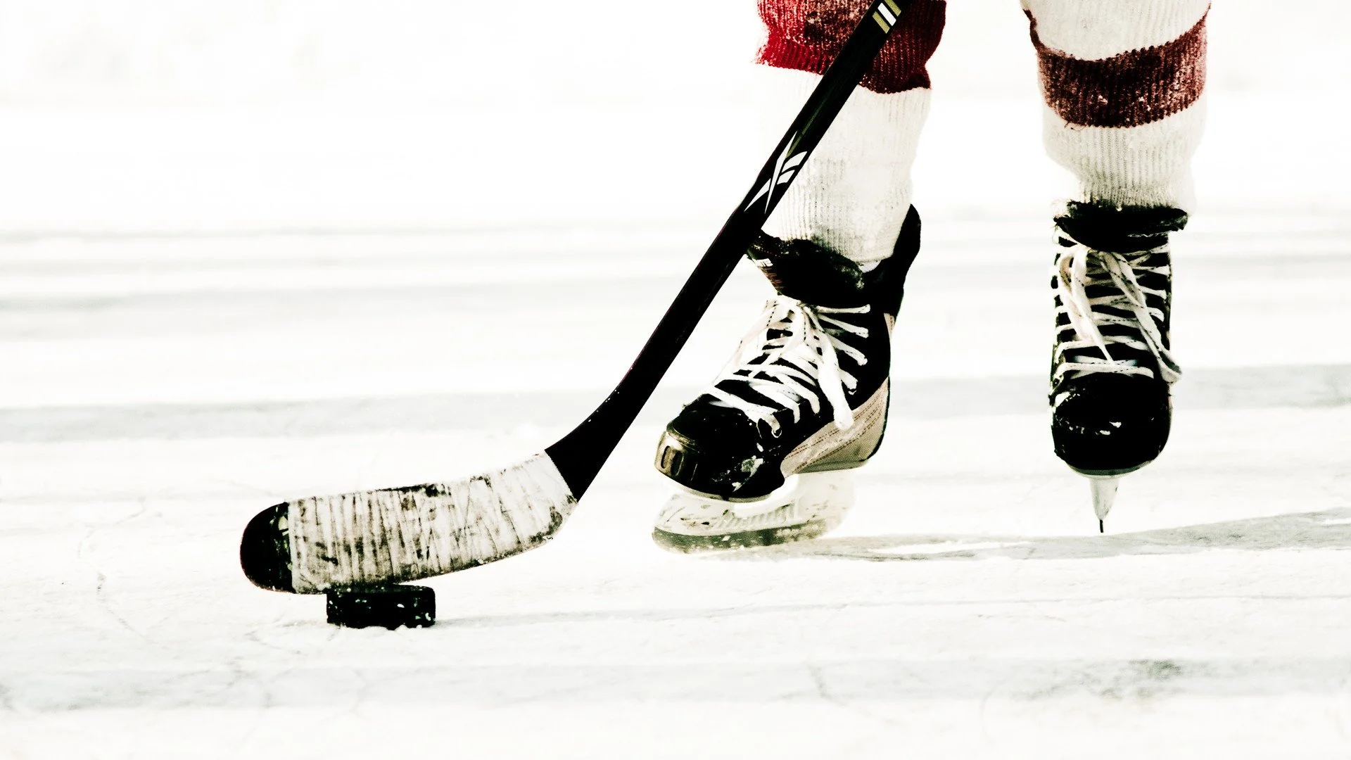 Close-up of ice hockey player snowboarding on ice, with hockey stick and puck, wearing black and white skates and striped socks.