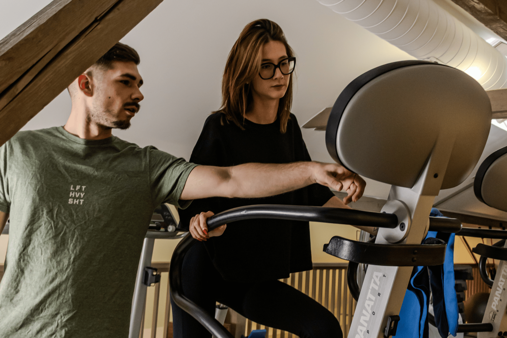 Zwei Menschen beim Training in einem Fitnessstudio, eine Frau sitzt auf einem Crosstrainer, ein Mann assistiert ihr.