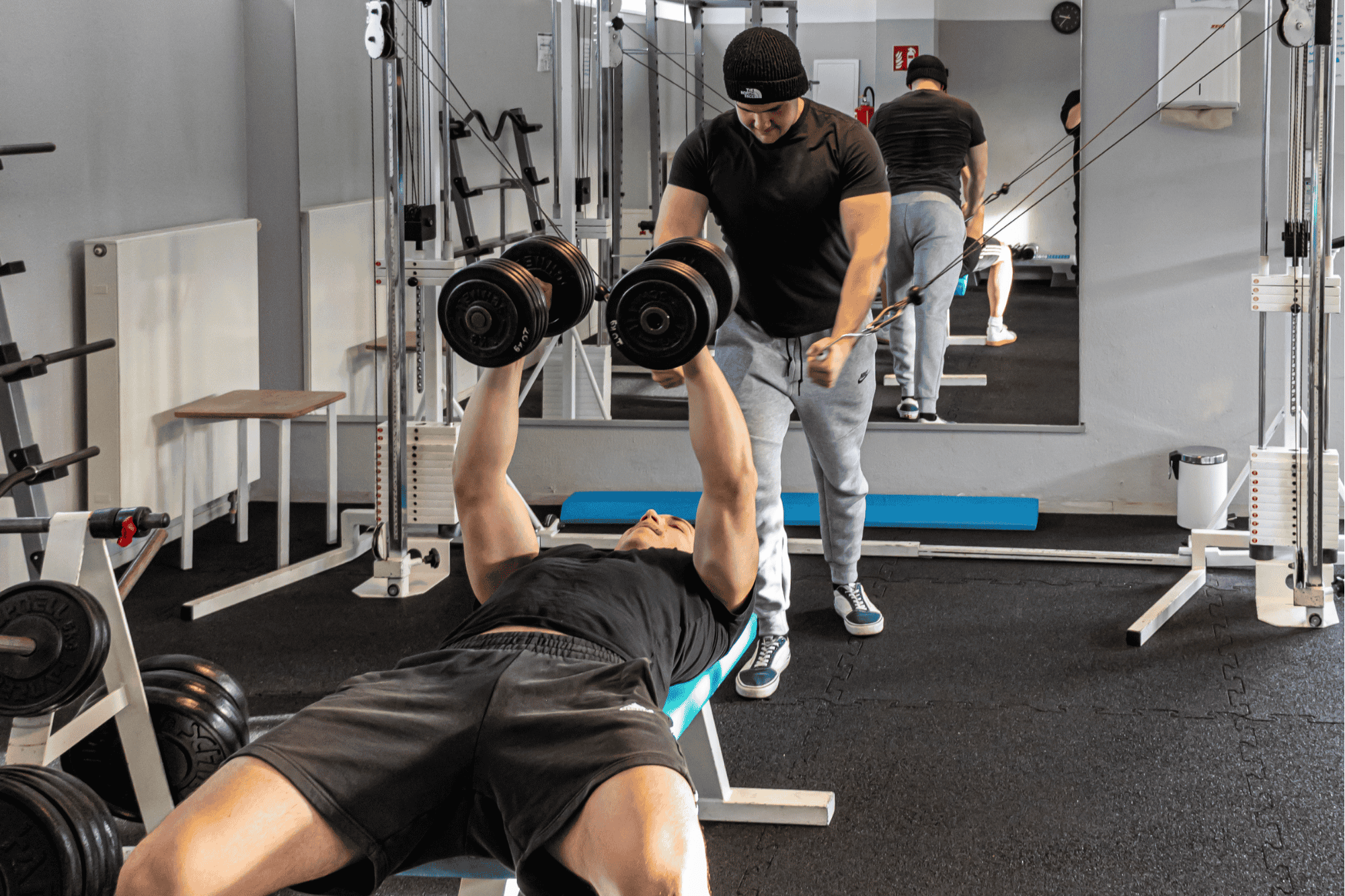 Ein Mann liegt auf einer Bank und hebt mit beiden Händen zwei Hanteln, während ein Trainer ihm einen Überblick gibt. Im Hintergrund sind zwei weitere Personen auf der Trainingsfläche.