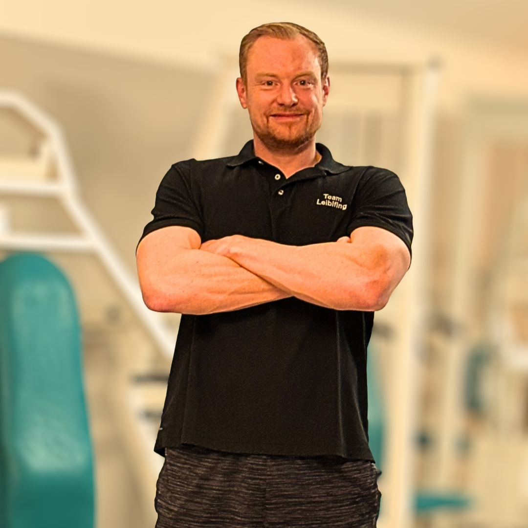 Ein Mann mit roten Haaren und Bart, der ein schwarzes Polo trägt mit dem Text "Toam Leibleing" auf der linken Brust, steht in einer Gymnasium-Umgebung, die im Hintergrund unscharf ist.