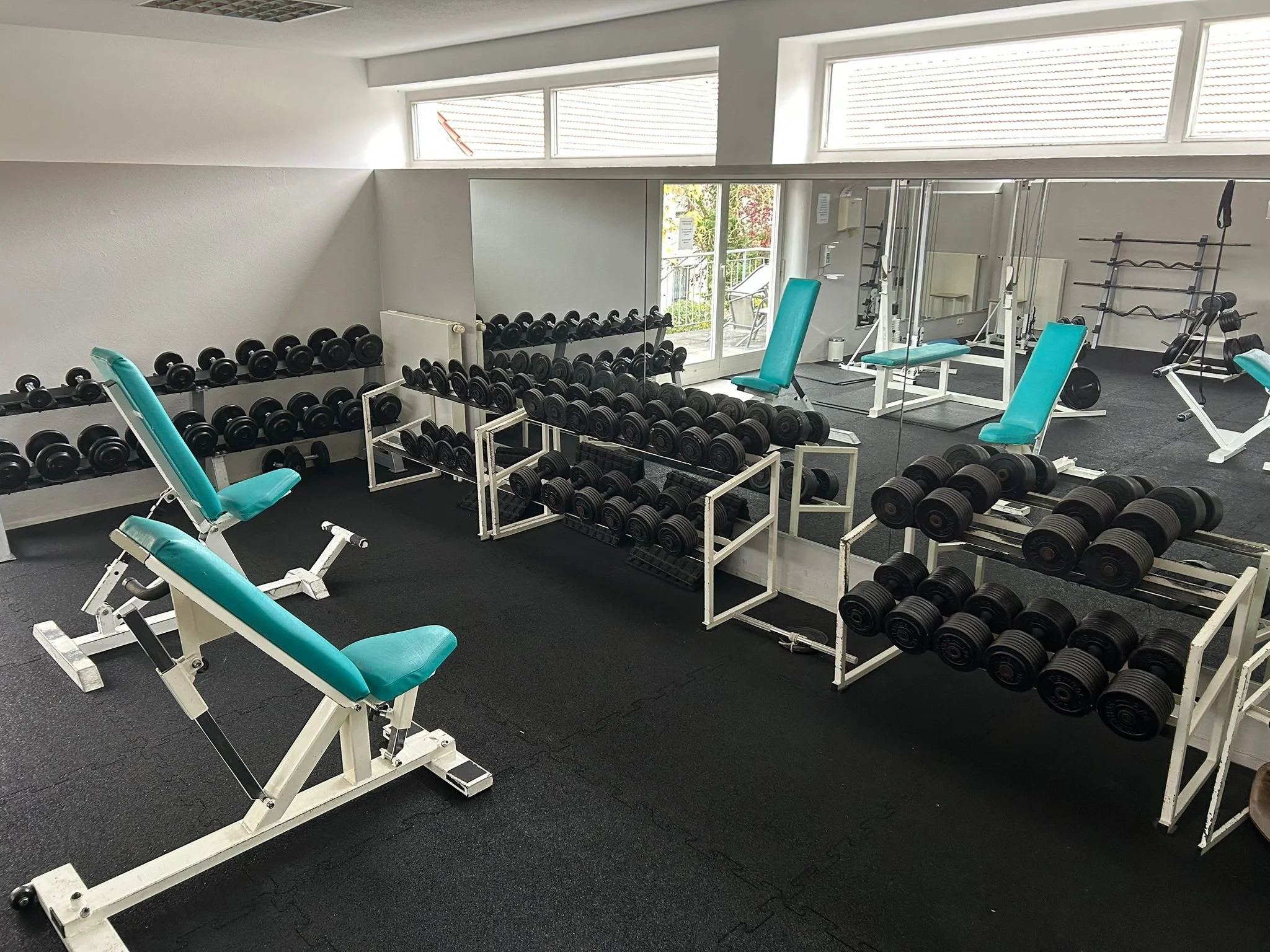 Ein aufgeräumtes Fitnessstudio mit Hanteln, Hantelrollständen, verstellbaren Trainingsbänken und Spiegeln an der Wand, gut beleuchtet durch große Fenster.