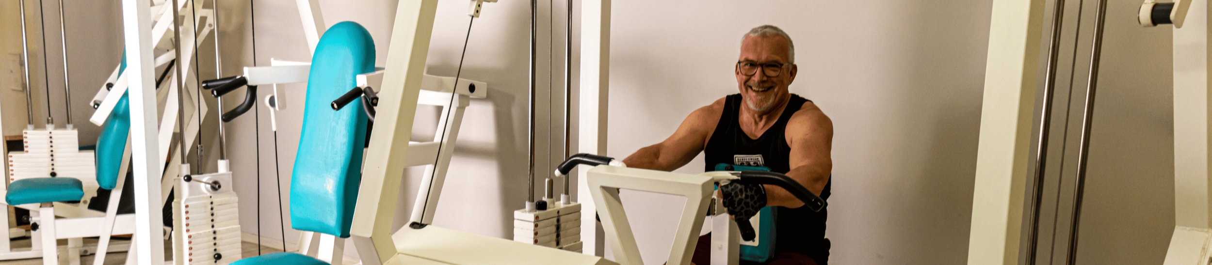 Ein lachender älterer Mann mit Brille und kurzen Haaren trainiert im Fitnessstudio mit einem Krafttrainingsgerät.