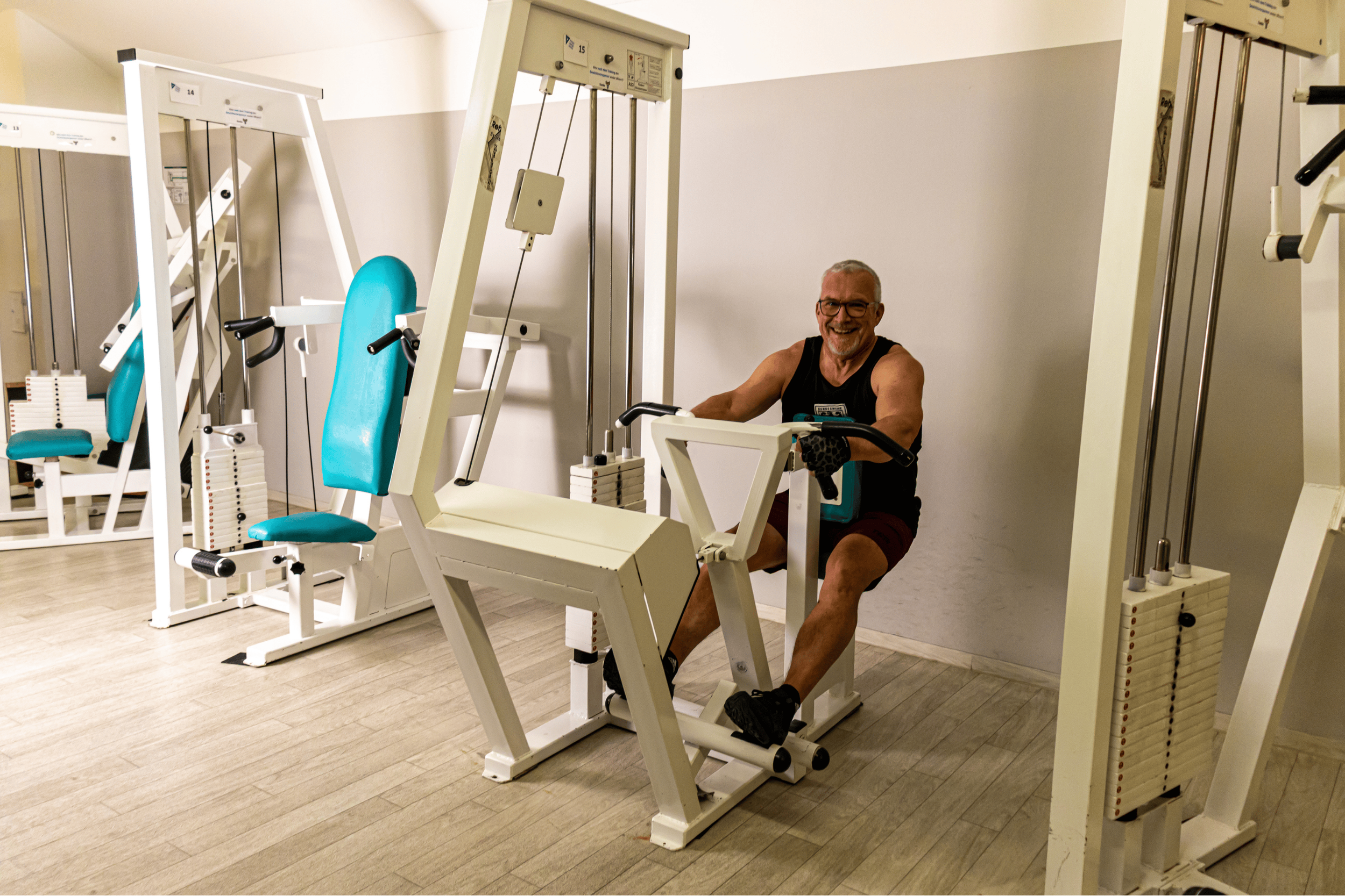 Ein Mann trainiert auf einem sitzenden Kraftgerät im Fitnessstudio, lächelt und trägt ein schwarzes Tanktop.