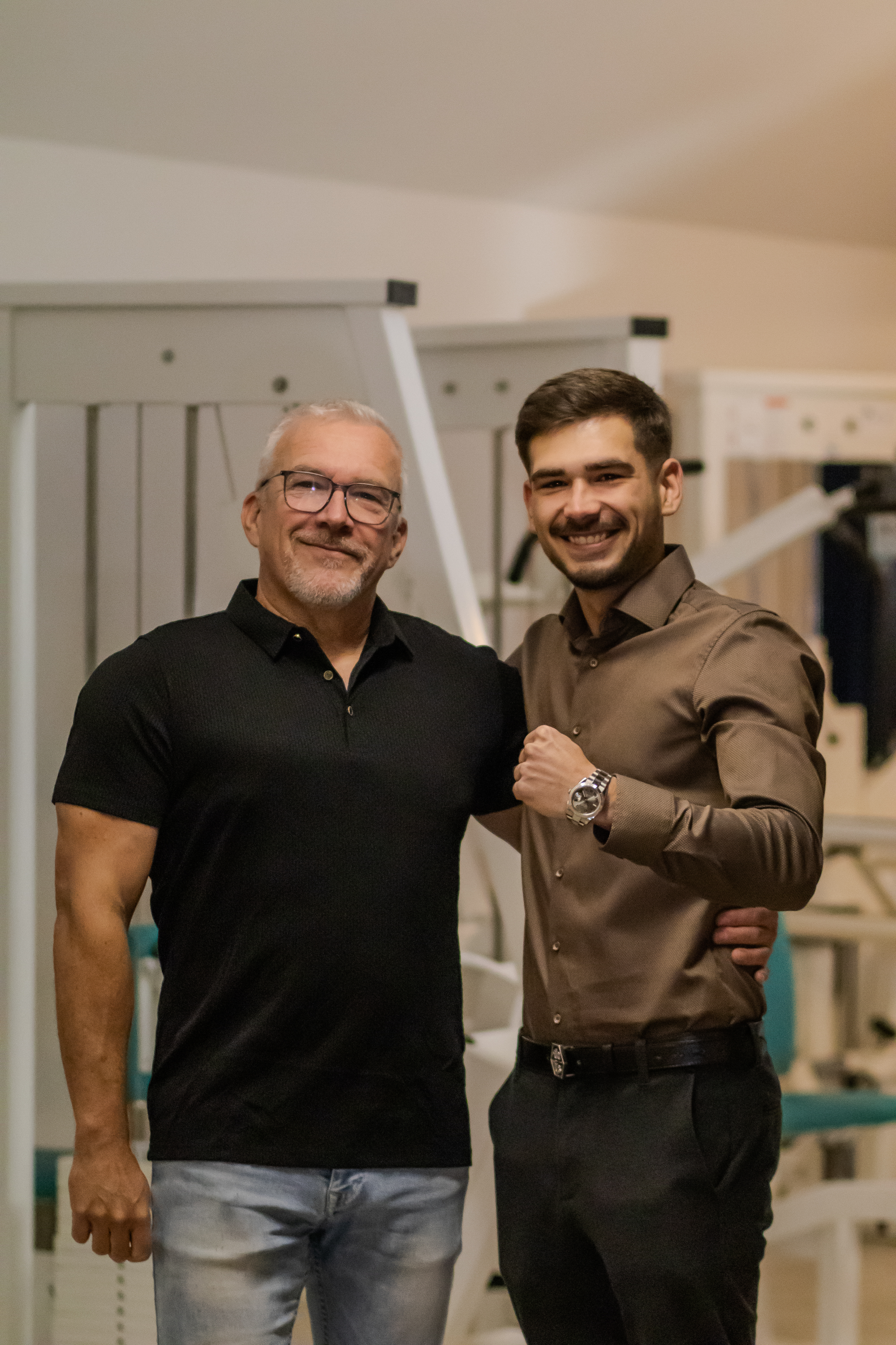 Zwei Männer stehen nebeneinander, lächelnd in einem Fitnessstudio.