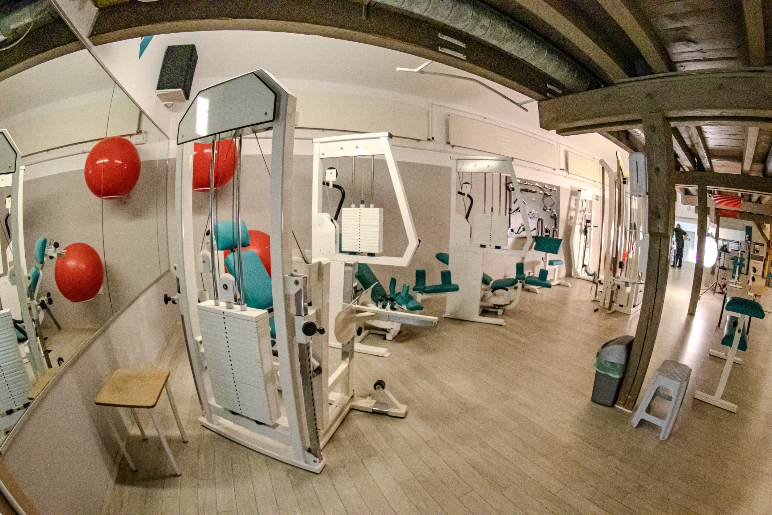 Fitnessstudio mit verschiedenen Trainingsgeräten und roten Bällen an der Wand, Holzfußboden, Spiegel an der Wand, Ecke mit Mülltonne und Hocker.