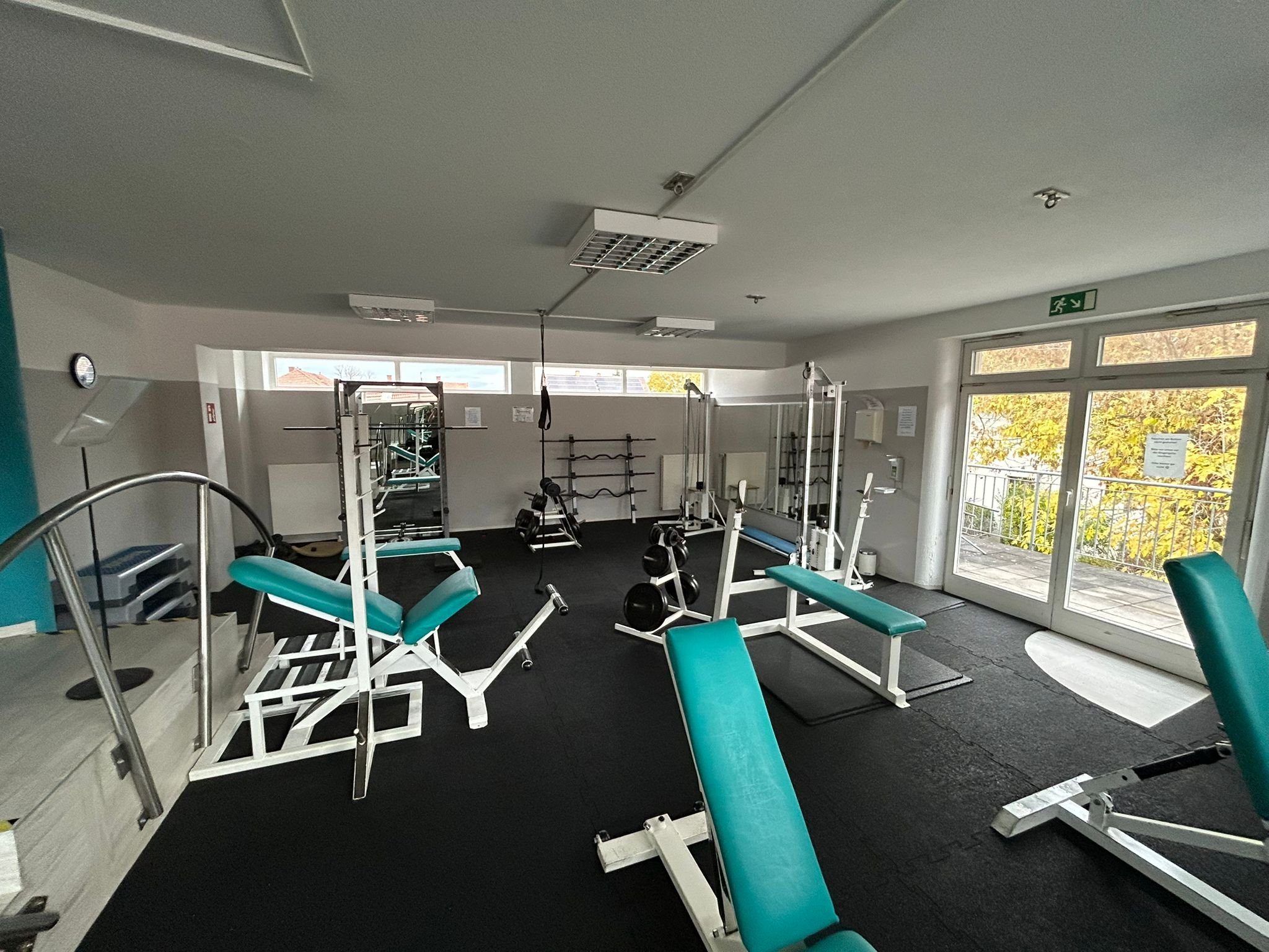 Ein Trainingsraum mit verschiedenen Fitnessgeräten, Hanteln, Hantelstangen, Hantelbänken, und Zugang zu einem Balkon mit Blick auf Bäume, beleuchtet durch Tageslicht.
