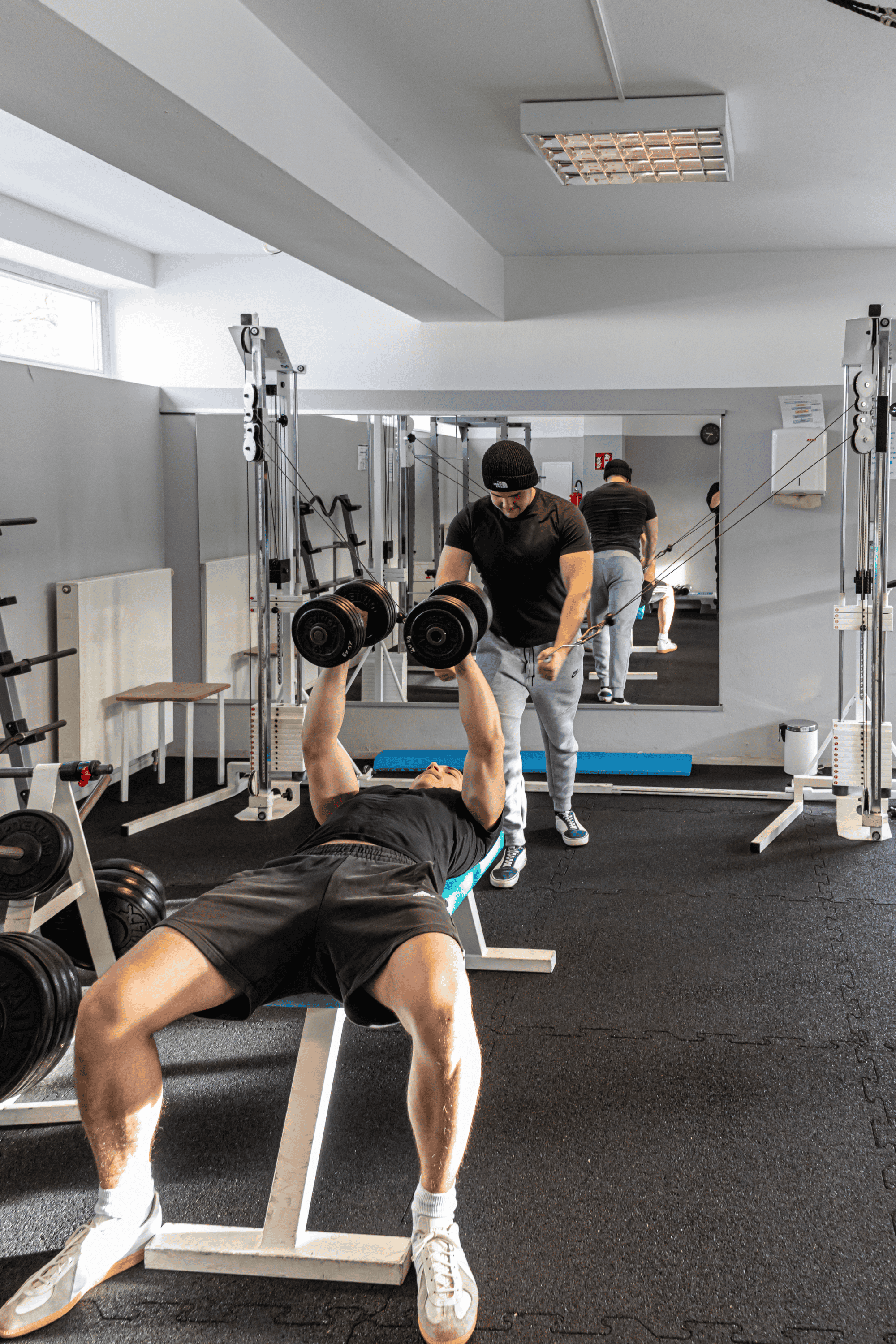 Person liegt auf einer Hantelbank und hebt mit beiden Händen Kurzhanteln, während ein Trainer neben ihm steht und ihn unterstützt. Im Hintergrund sind weitere Personen beim Training.