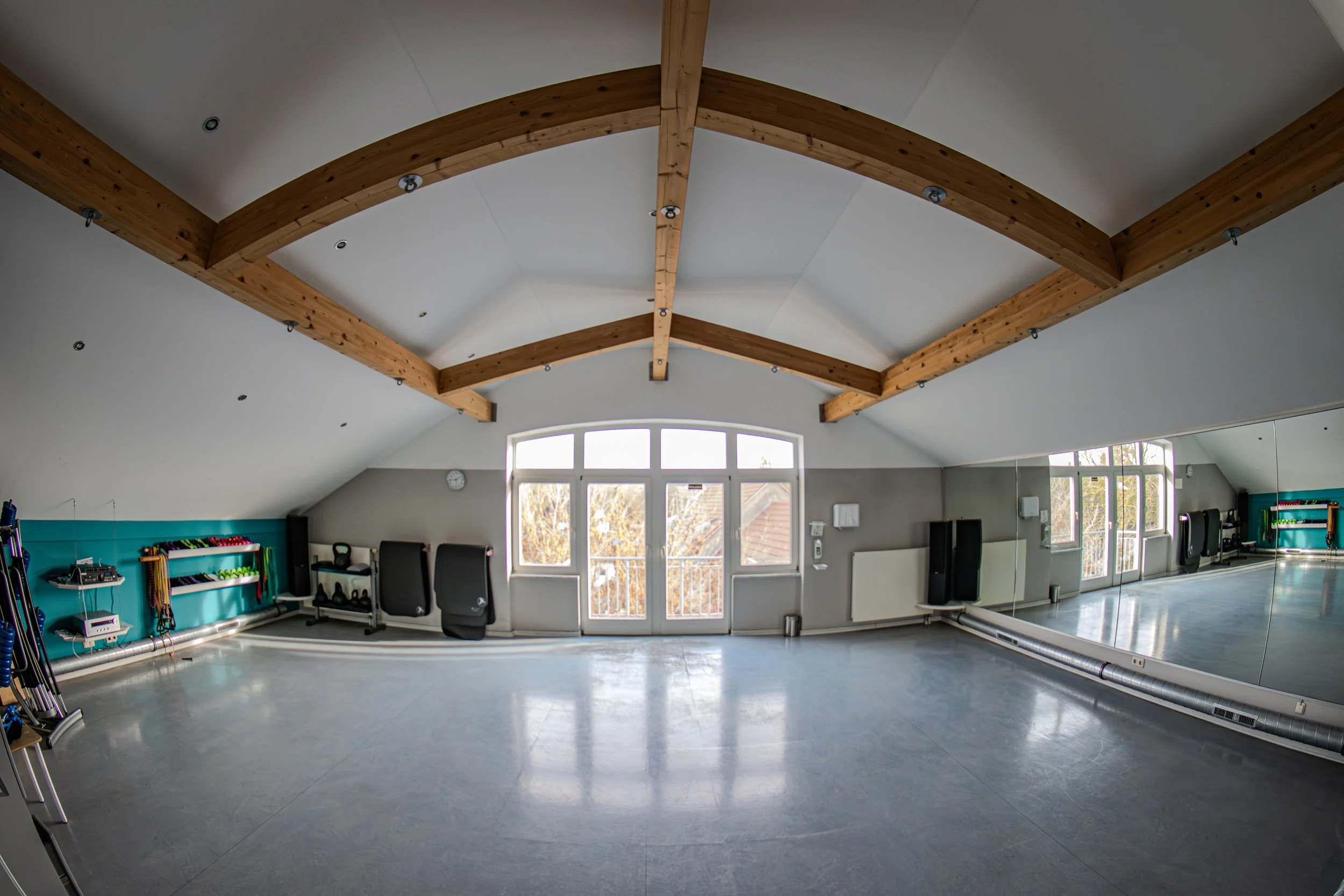 Ein großer, heller Fitnessraum mit einer hohen Decke und sichtbaren Holzbalken. Der Raum hat große Fenster, die natürliches Licht hereinlassen, und eine Spiegelwand auf der rechten Seite. Fitnessgeräte wie Hanteln, Widerstandsband-Aktionen und ein pa