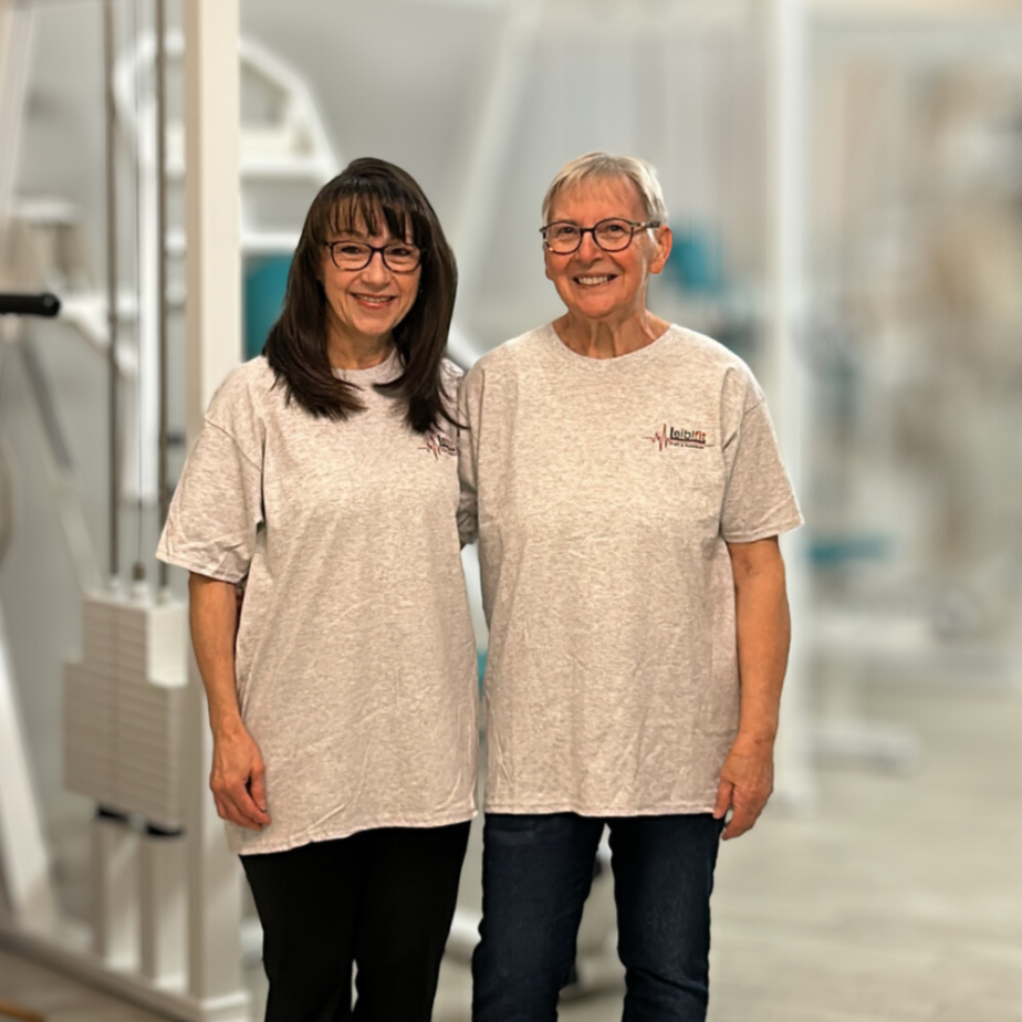 Zwei Frauen stehen zusammen in einem Fitnessstudio, beide tragen graue T-Shirts und lächeln.