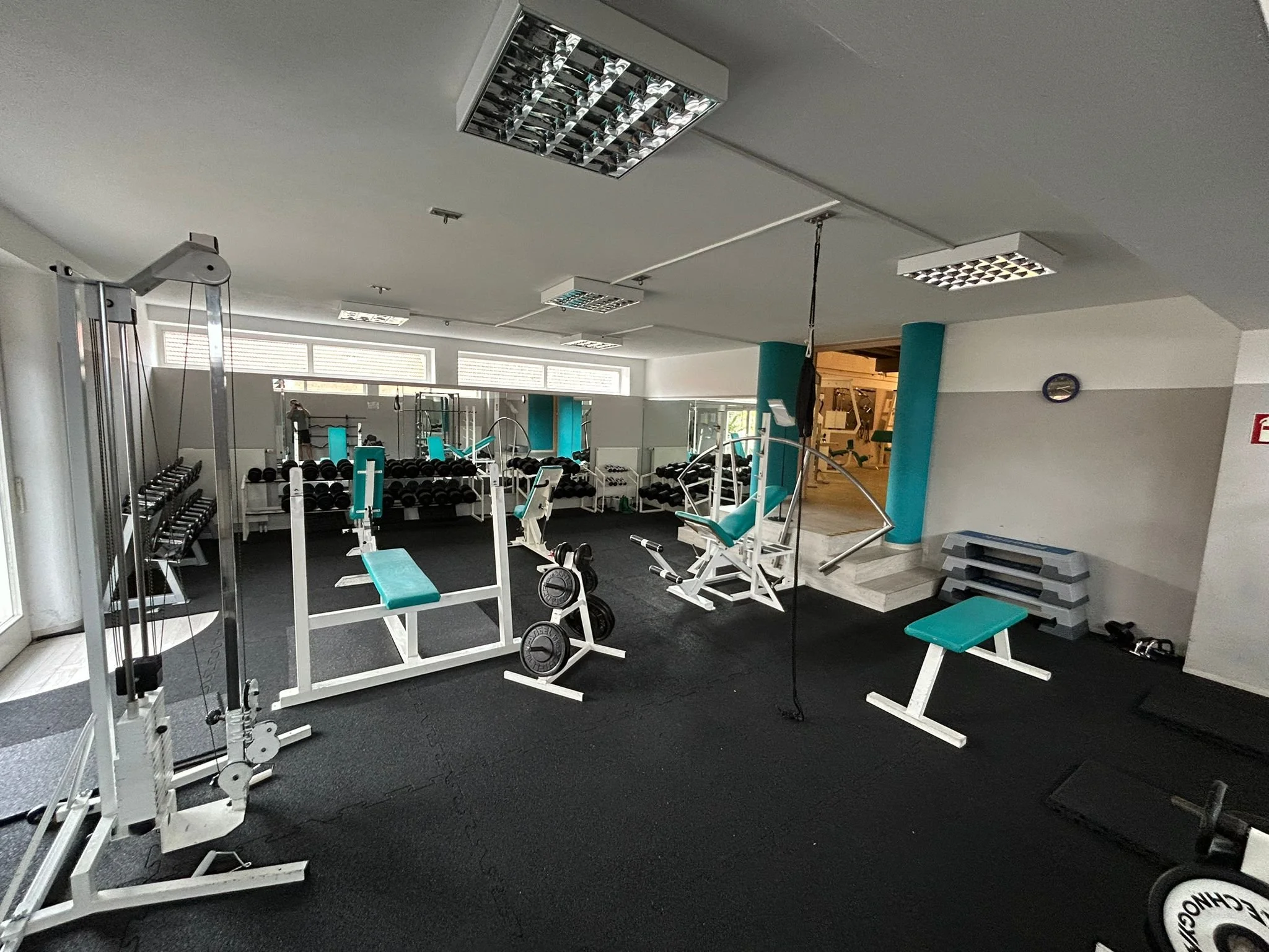 Innenraum eines Fitnessstudios mit verschiedenen Trainingsgeräten, Hanteln, Steppern und Fitnessbänken, gut beleuchtet durch große Fenster.