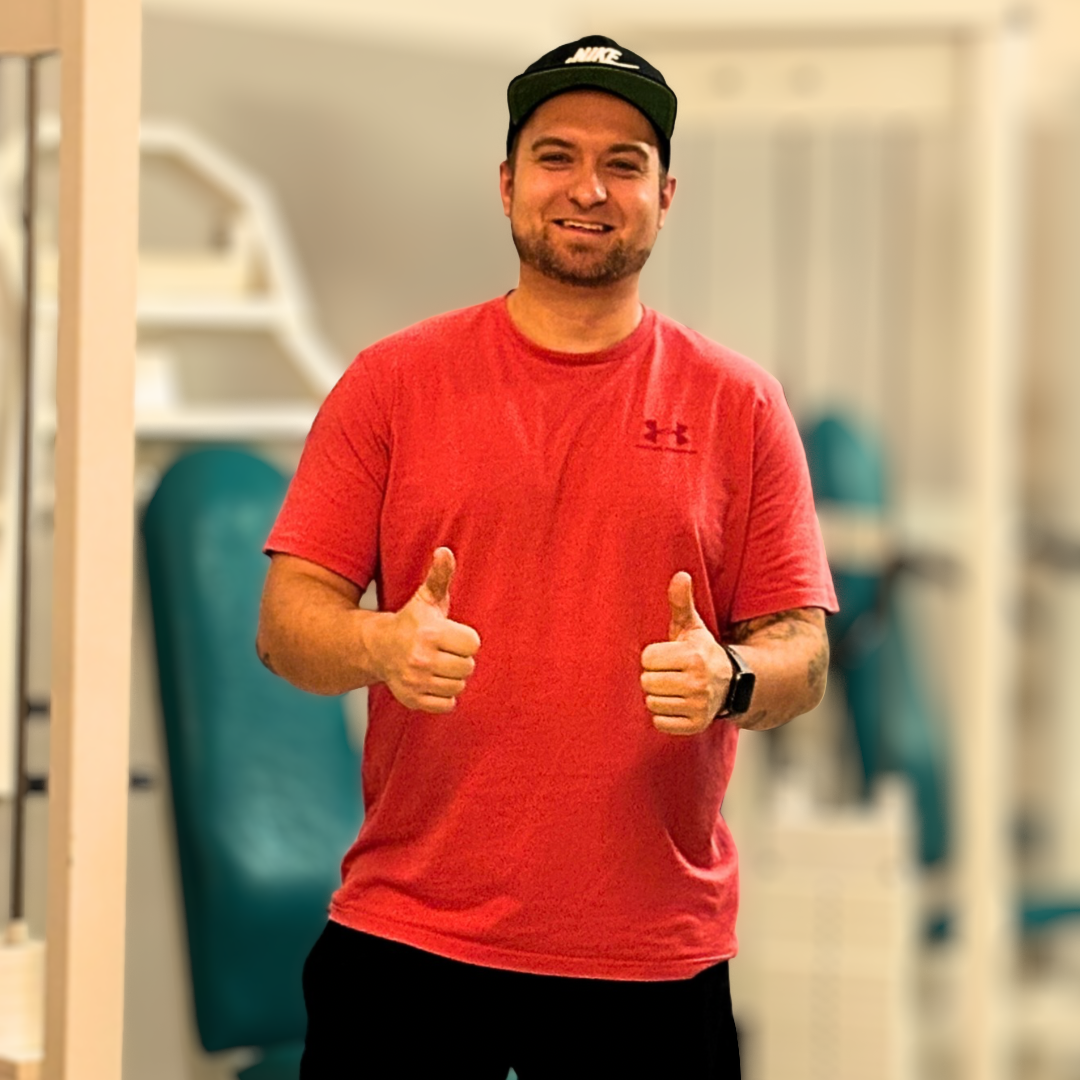 Mann in rotem T-Shirt, schwarzem Cap und Smartwatch, der beide Daumen hoch zeigt und lächelt, im Fitnessstudio.
