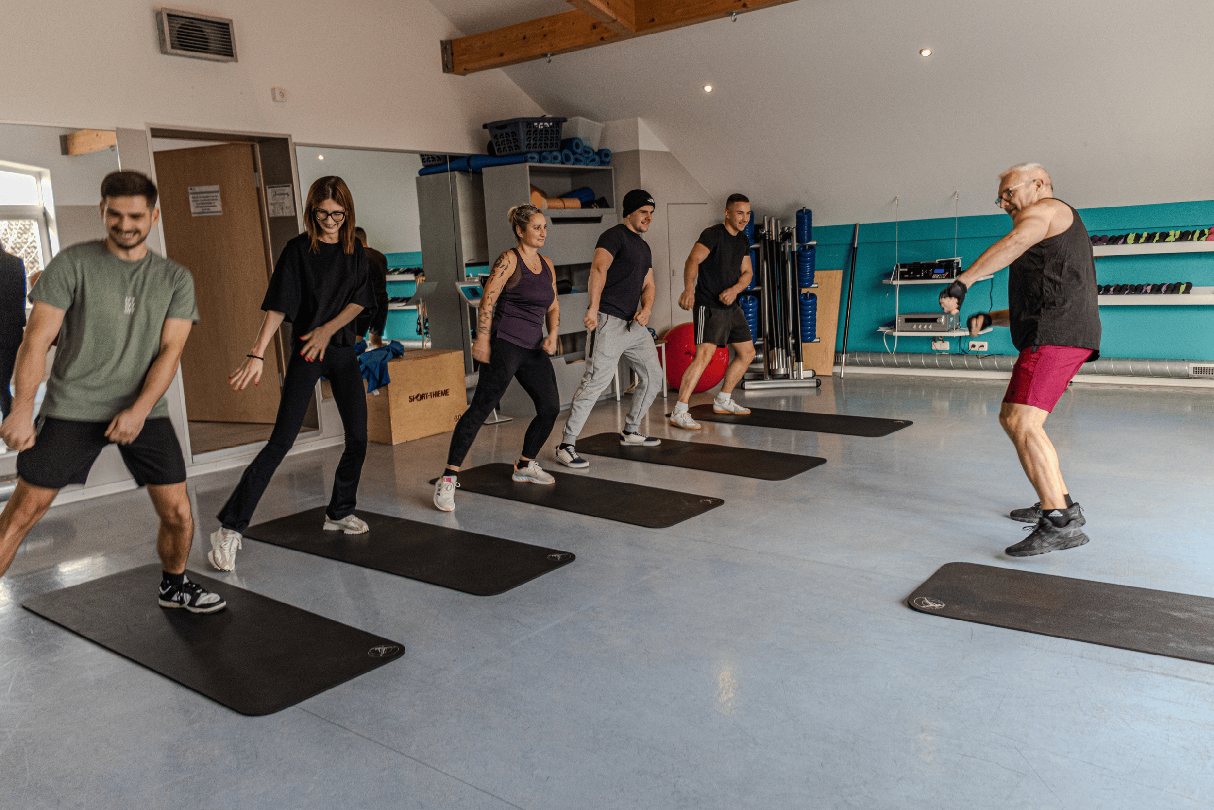Gruppe von Menschen, die an einem Fitnesskurs in einem Fitnessstudio teilnehmen, alle stehen auf Gymnastikmatten und folgen einem Trainer, der Übungen macht.