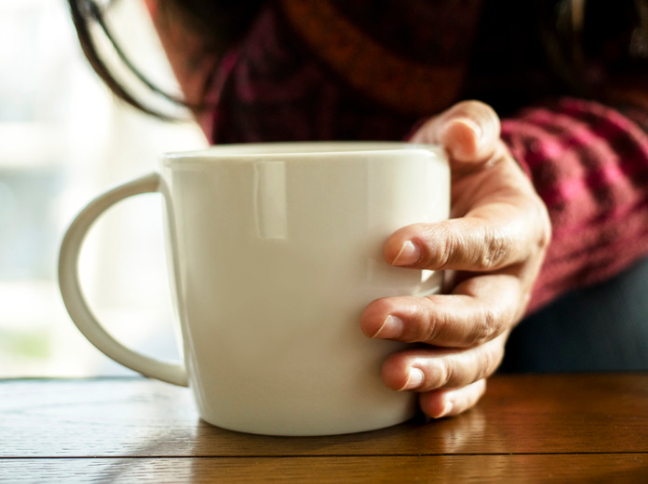 A person holding a white ceramic coffee mug with their left hand, sitting at a wooden table.