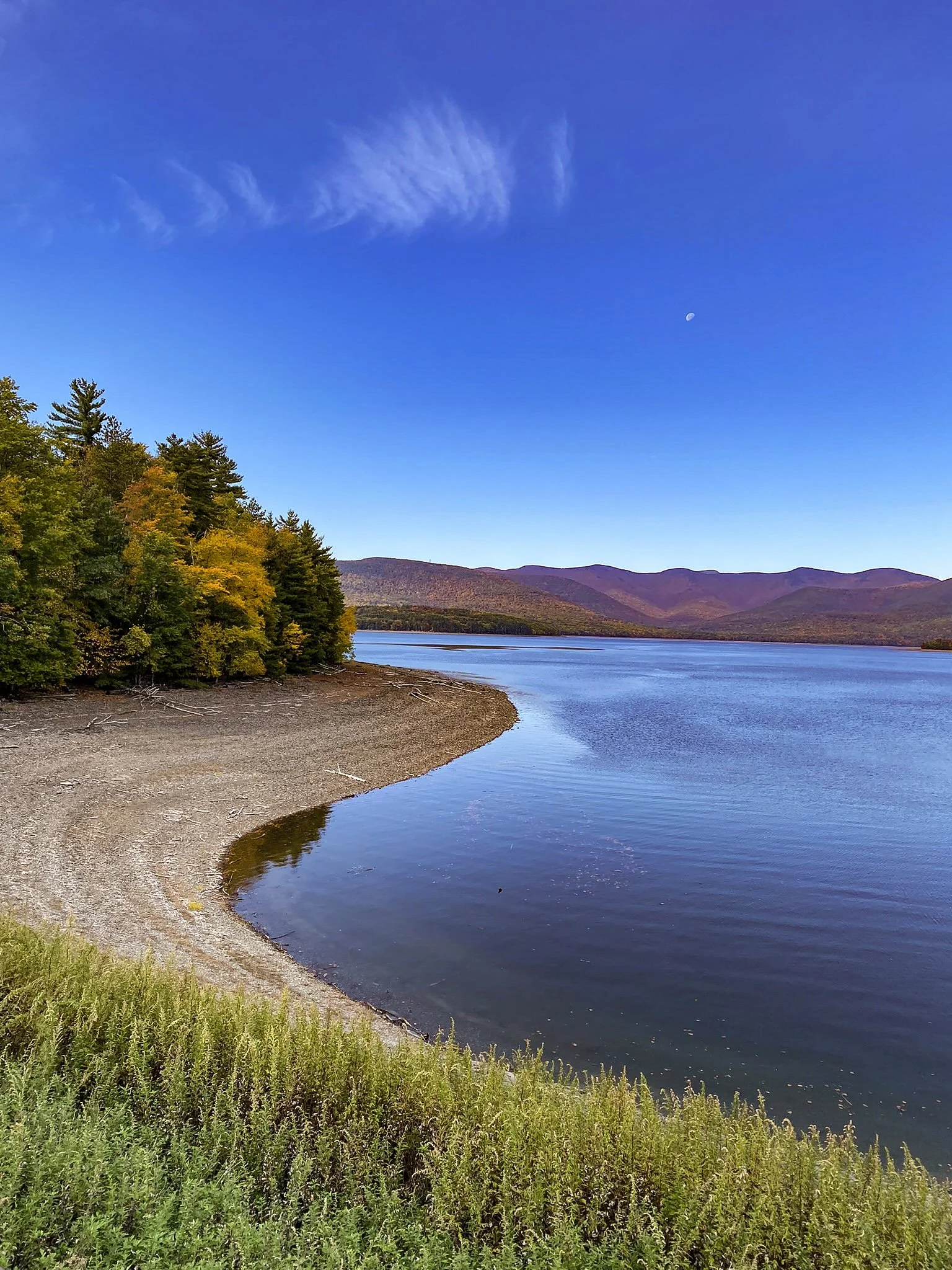 A peaceful lake surrounded by trees with vibrant autumn foliage, a rocky shore, and distant mountains under a clear blue sky with wispy clouds and a visible moon.